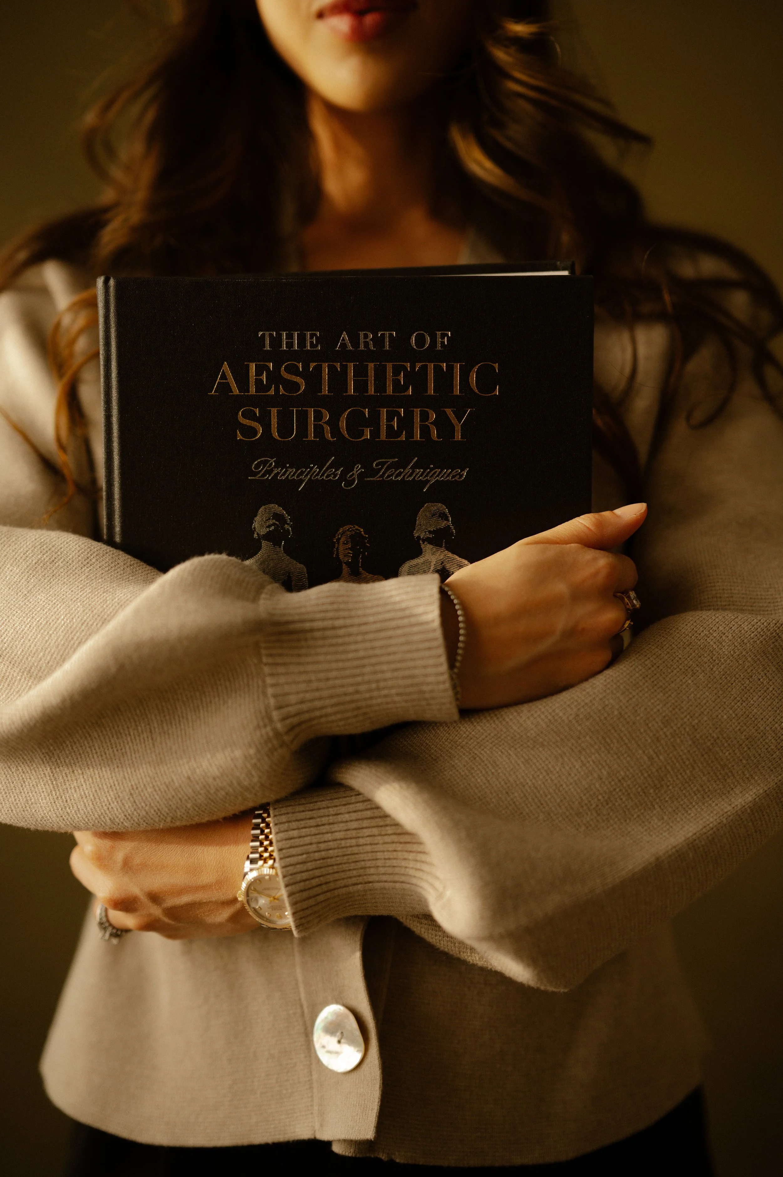 A person holding a black book titled "The Art of Aesthetic Surgery Principles & Techniques" while wearing a beige sweater, a watch, and rings, with long wavy hair and a blurred background.