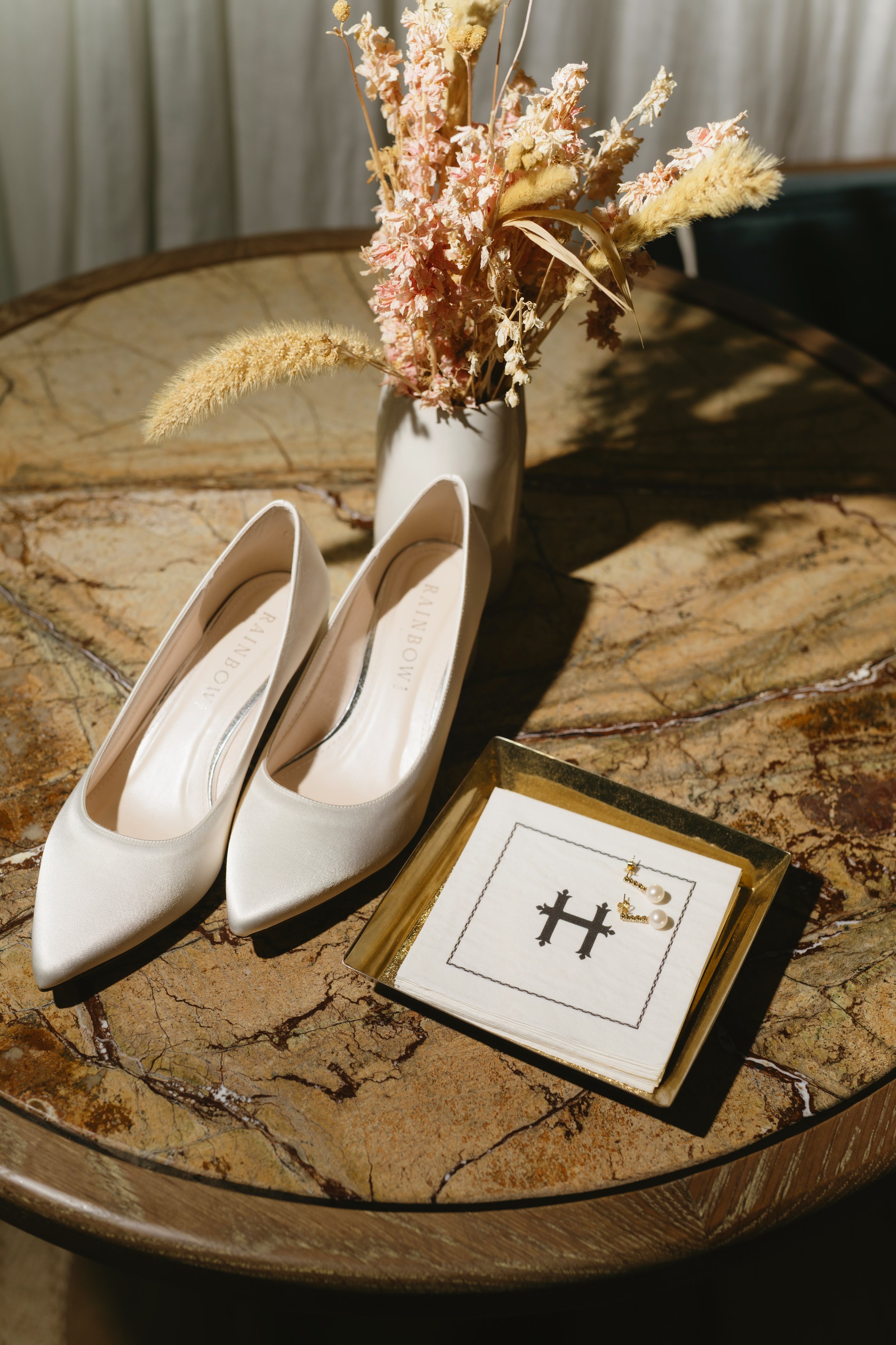 A pair of white high-heeled shoes with large round rhinestone brooch embellishments, a gold wedding band, an engagement ring with a large center stone, and a diamond eternity band, all arranged on a white surface.