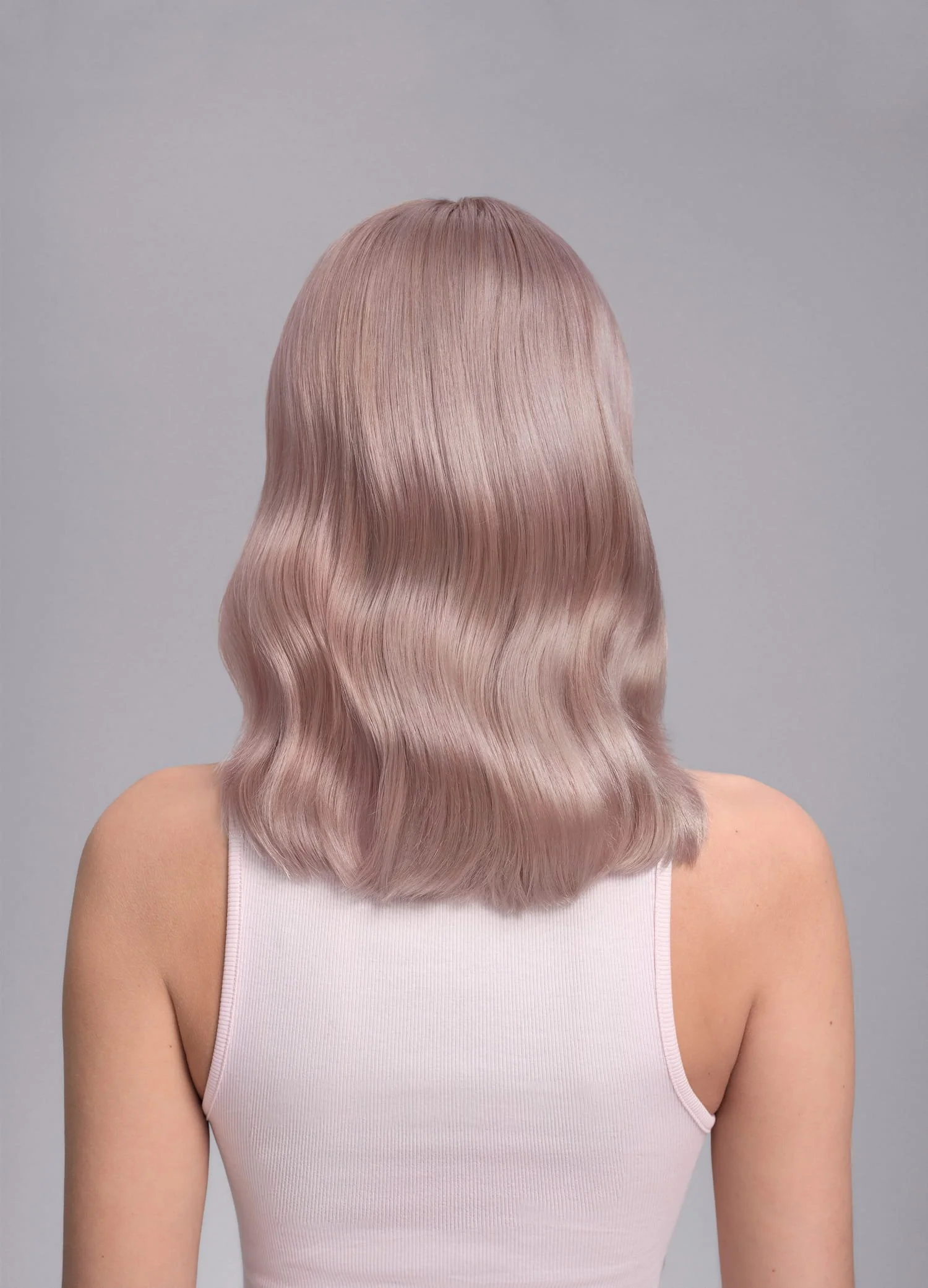 Back view of a woman with shoulder-length, wavy, pastel pink hair, wearing a white tank top against a gray background.