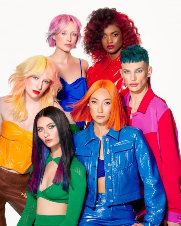 Six women with colorful hairstyles and outfits posing together against a white background.
