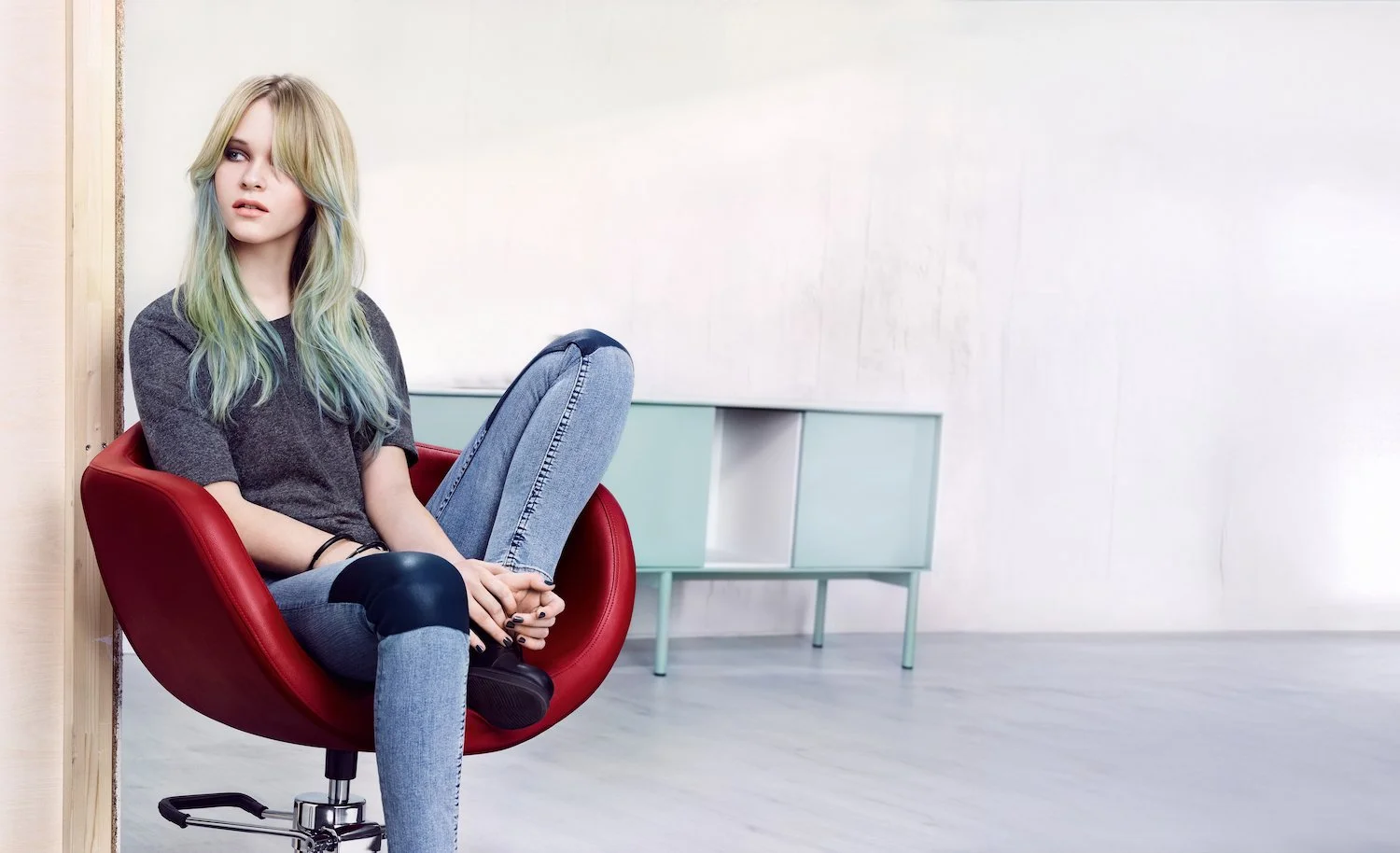 A young woman with long, wavy, pastel-colored hair sitting in a modern red chair in a minimalist room with white walls and a light green storage unit in the background.