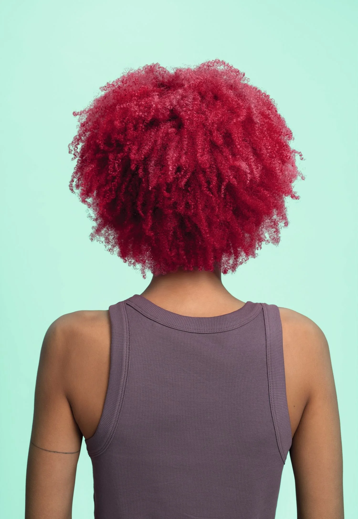 Back of a person with vibrant red, curly, natural hair, wearing a purple sleeveless top against a light turquoise background.