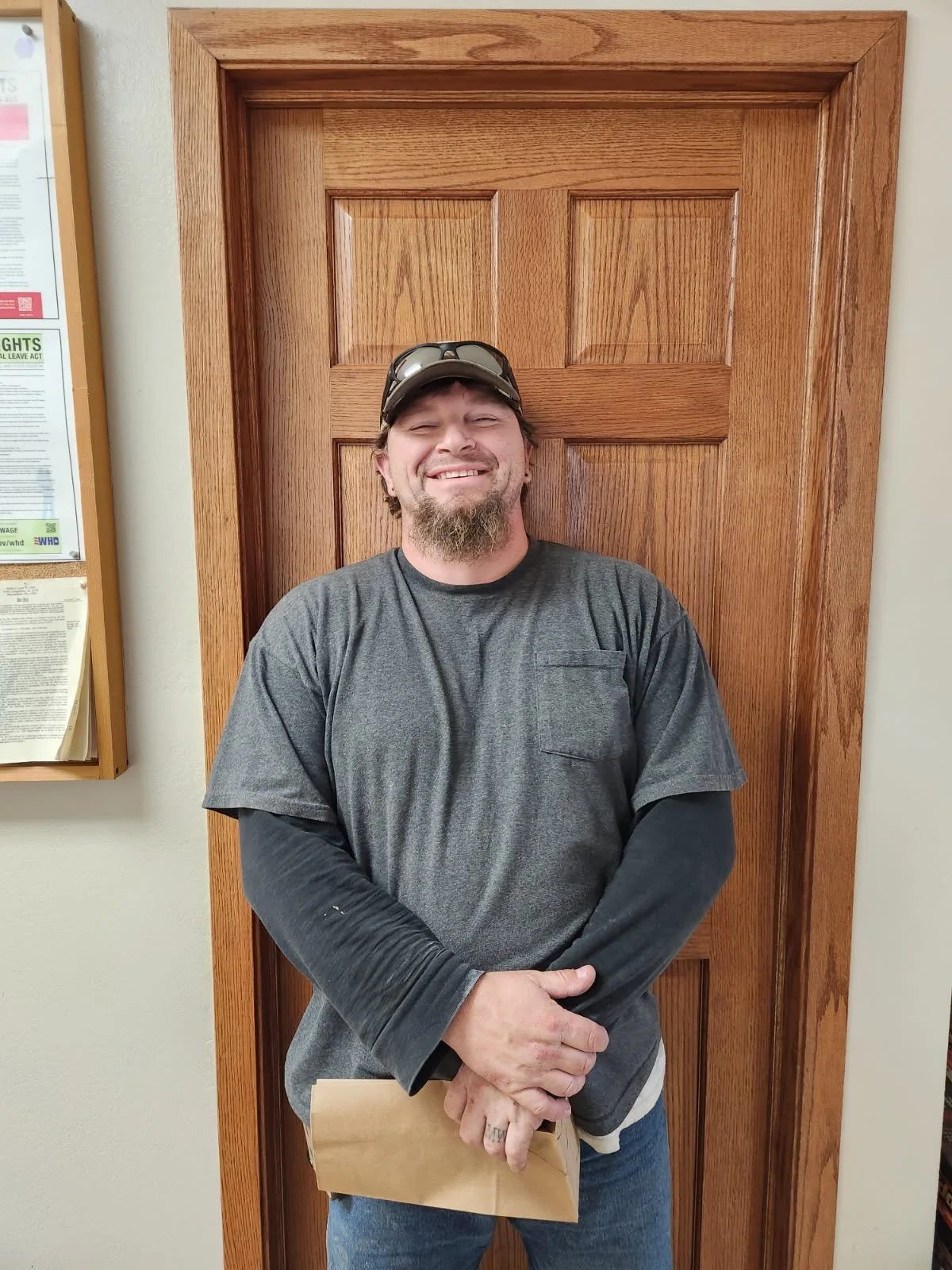 Dan Wrobleski. A man with a beard, sunglasses on his head, wearing a dark gray T-shirt over a long-sleeve black shirt, standing in front of a wooden door, smiling with his hands clasped, holding a paper envelope.
