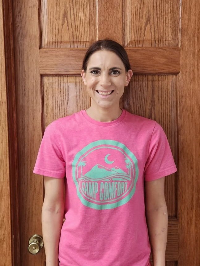 Shawna Cross. A woman standing in front of a wooden door, smiling, wearing a pink t-shirt with a graphic that says 'Camp Comfort' and features a mountain, palm trees, and a crescent moon.
