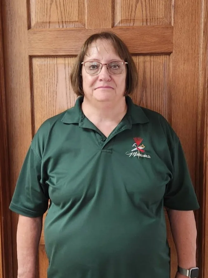 Cindy Seppala. An older woman with short brown hair, glasses, and a neutral expression standing in front of a wooden door. She is wearing a dark green polo shirt with a logo of a bird and red feathers on the left side.