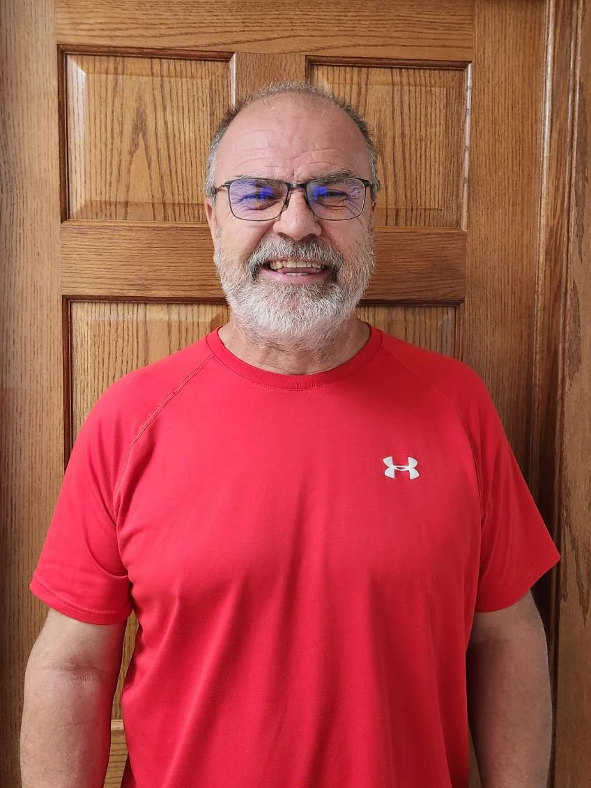 John Friman. A man with glasses, a beard, and gray hair smiling, standing in front of a wooden door, wearing a red Under Armour t-shirt.