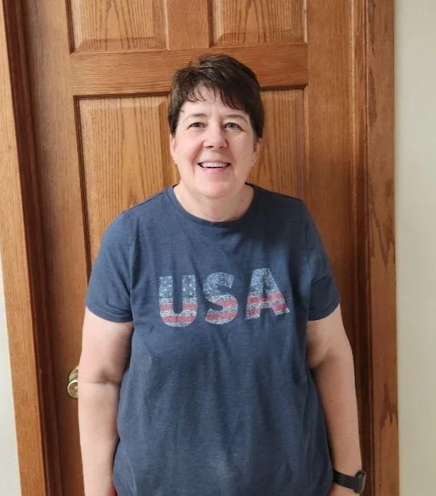 Michelle White. A smiling woman with short dark hair wearing a navy blue T-shirt with a faded USA flag design standing in front of a wooden door.