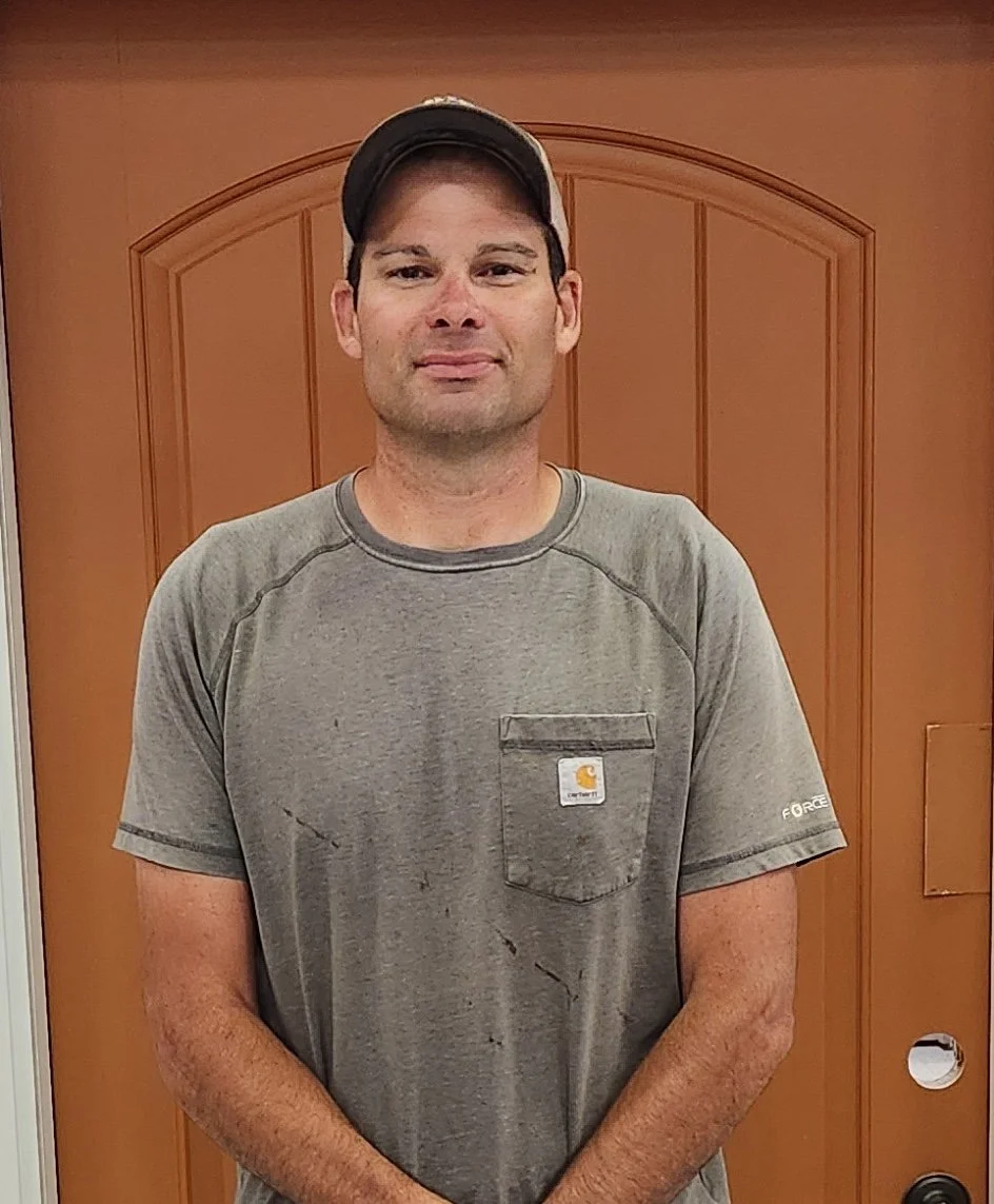 Dan Brunskill. A young man wearing a gray Carhartt T-shirt and a gray baseball cap, standing indoors in front of a wooden door.