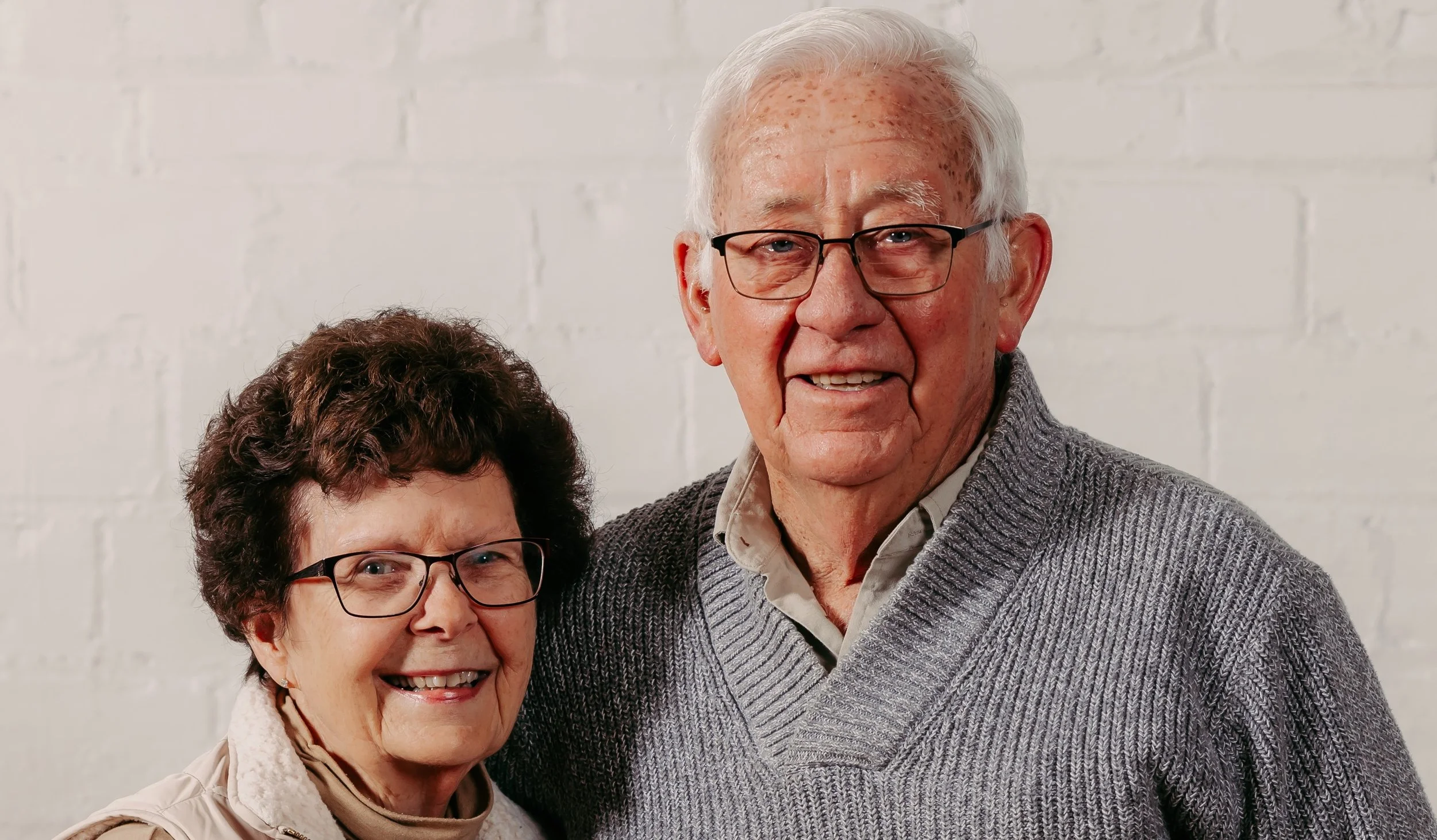 Bob and Gail Mueller, Founders. An elderly couple smiling together against a white brick wall. The woman has curly brown hair and glasses, wearing a cream jacket. The man with white hair and glasses, wearing a gray sweater and a collared shirt.