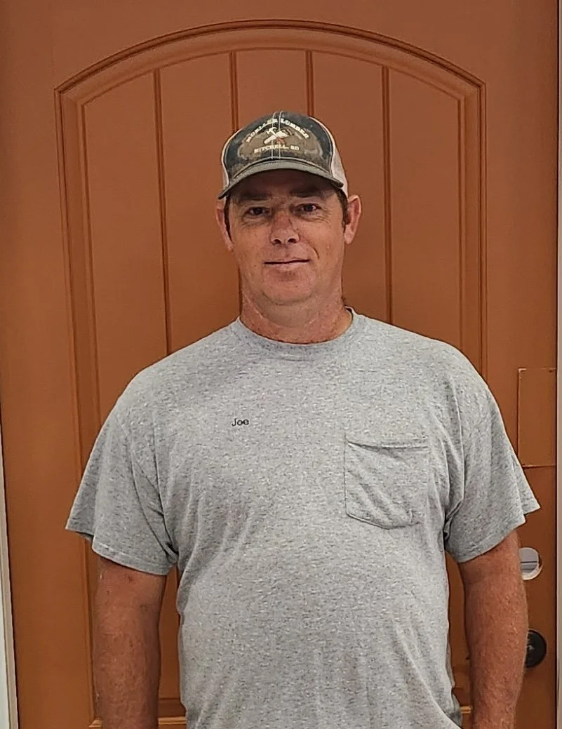 Joe Royston. A man wearing a grey t-shirt and a baseball cap standing in front of a wooden door.