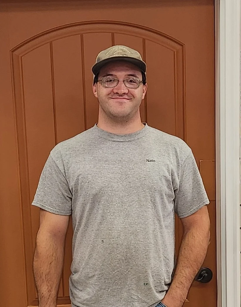 Nate Zirpel. A man wearing a grey t-shirt, glasses, and a beige cap standing indoors in front of a wooden door, smiling at the camera.