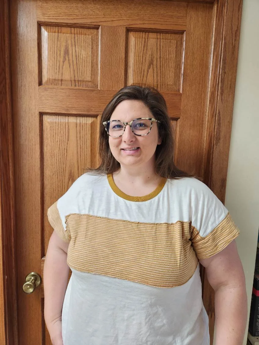 Skye Gaulke. A woman with brown hair, glasses, and light skin standing in front of a wooden door, wearing a white t-shirt with a tan and gold striped panel across the chest.