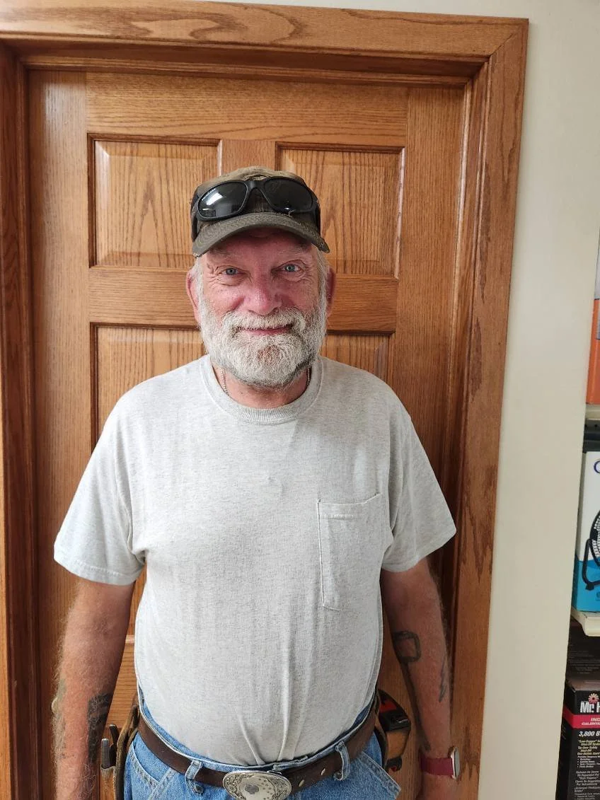 Paul Weichart. A smiling older man with a gray beard, wearing a light gray t-shirt, a baseball cap with sunglasses on top, and a belt with a large belt buckle, standing in front of a wooden door.