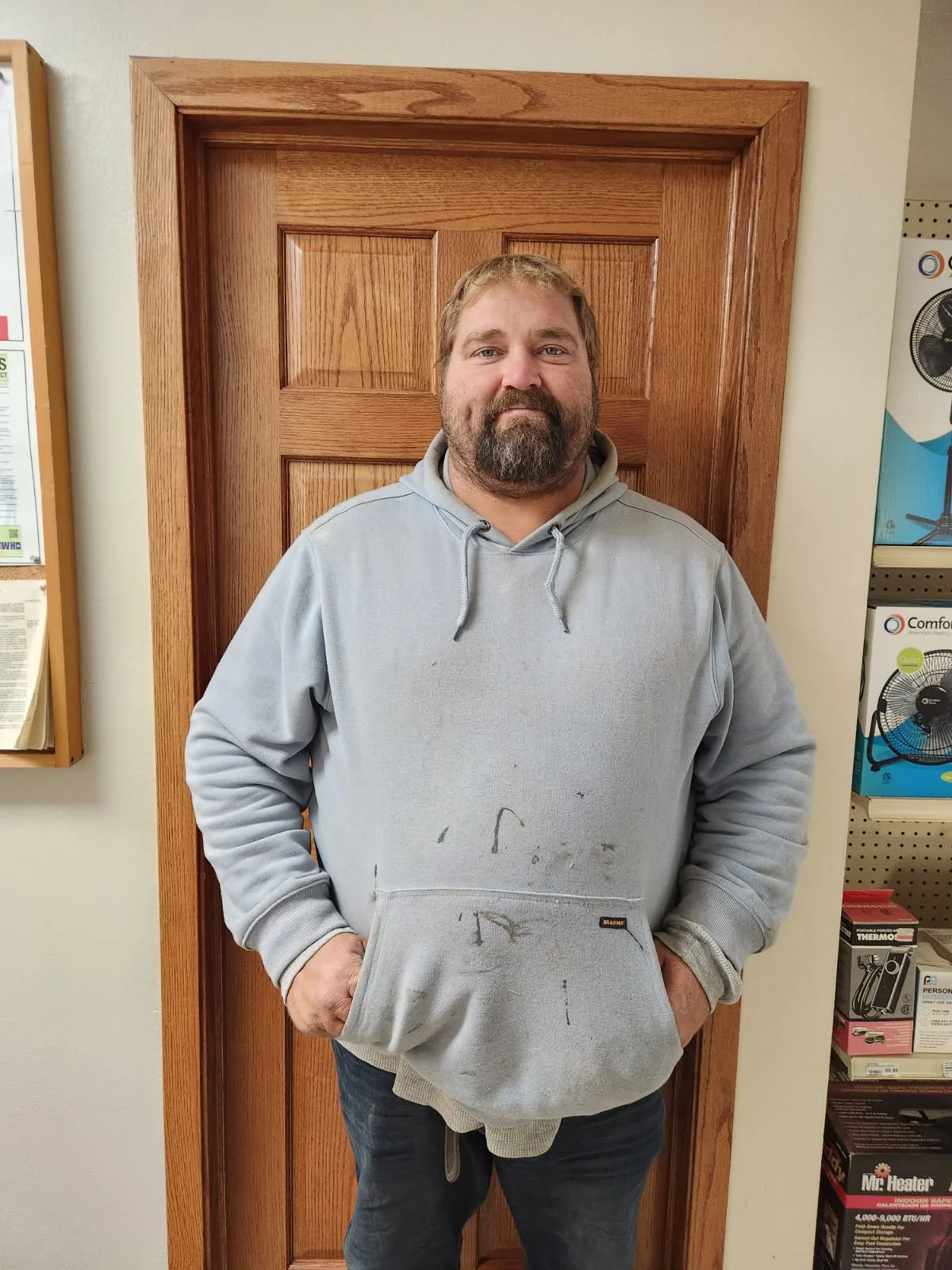 Stacy Maeschen. A man with a beard and light brown hair standing in front of a wooden door. He is wearing a light gray hoodie with stains and has his hands in the hoodie's pocket.