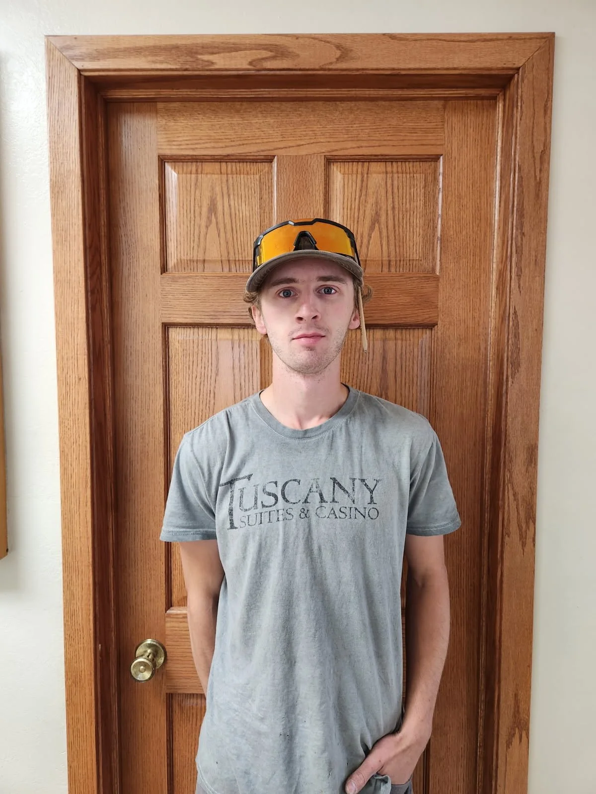 Dylan Reimnitz. Young man standing in front of a wooden door, wearing a gray T-shirt with "Tuscany Suites & Casino" printed on it, and a gray cap with goggles on top.