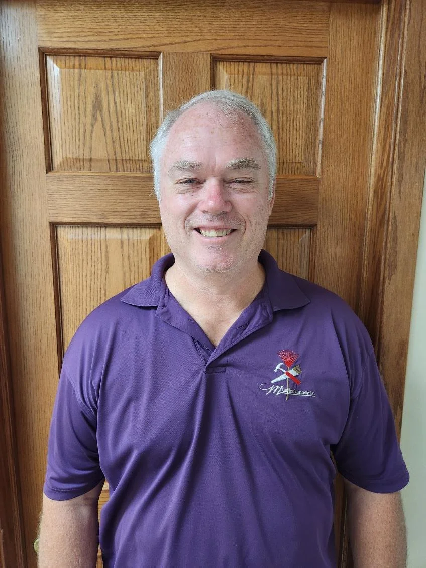 Mark White. A smiling middle-aged man with gray hair wearing a purple collared shirt standing in front of a wooden door.