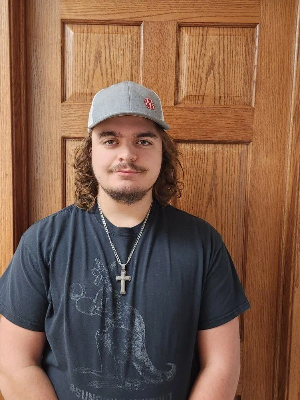 Jackson Roy. Young man with curly brown hair and facial hair wearing a gray baseball cap, a black T-shirt with a dinosaur graphic, and a cross necklace, standing in front of a wooden door.