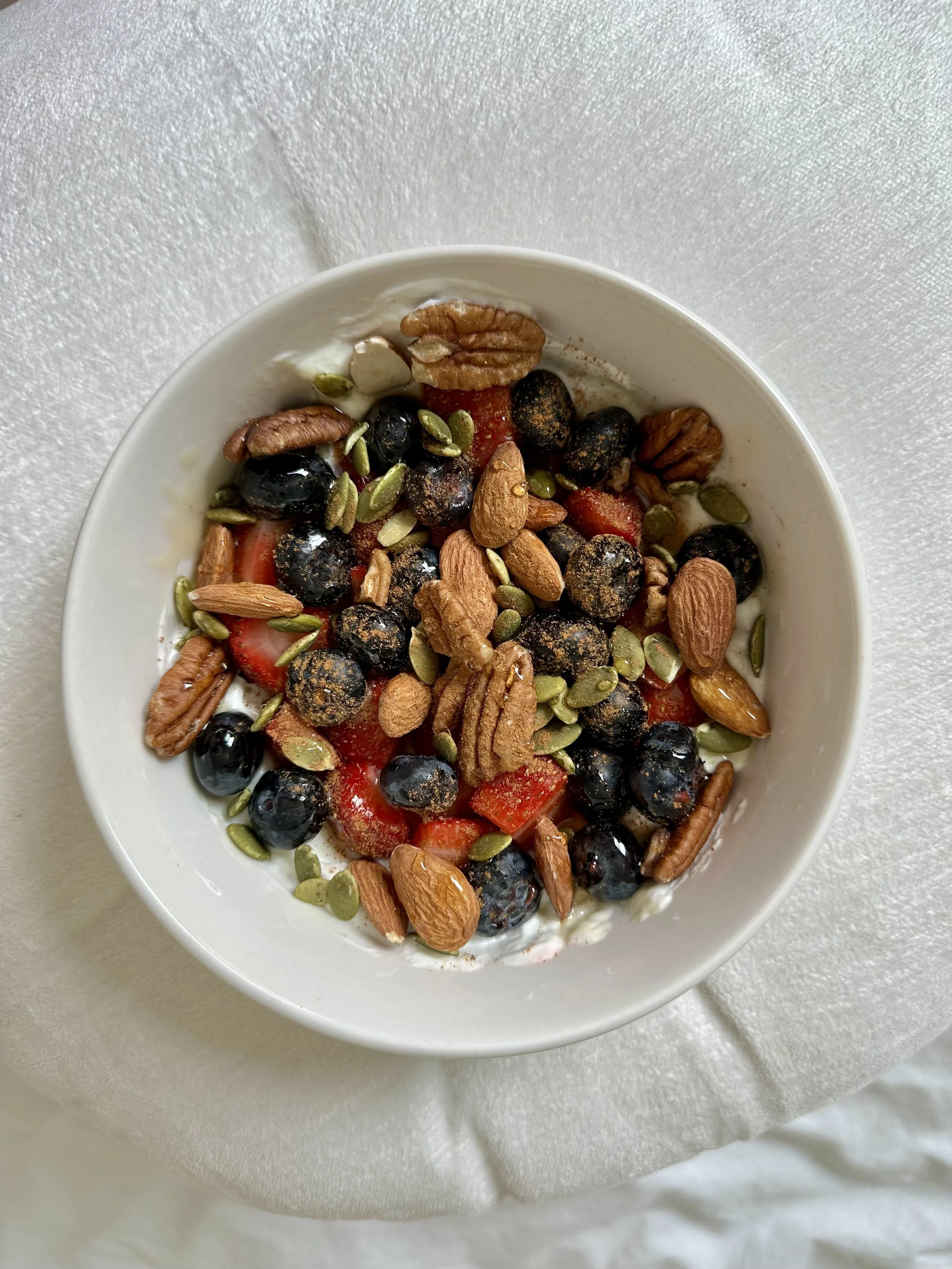 A bowl of yogurt topped with blueberries, strawberries, nuts, pumpkin seeds, and almonds on a white textured surface.