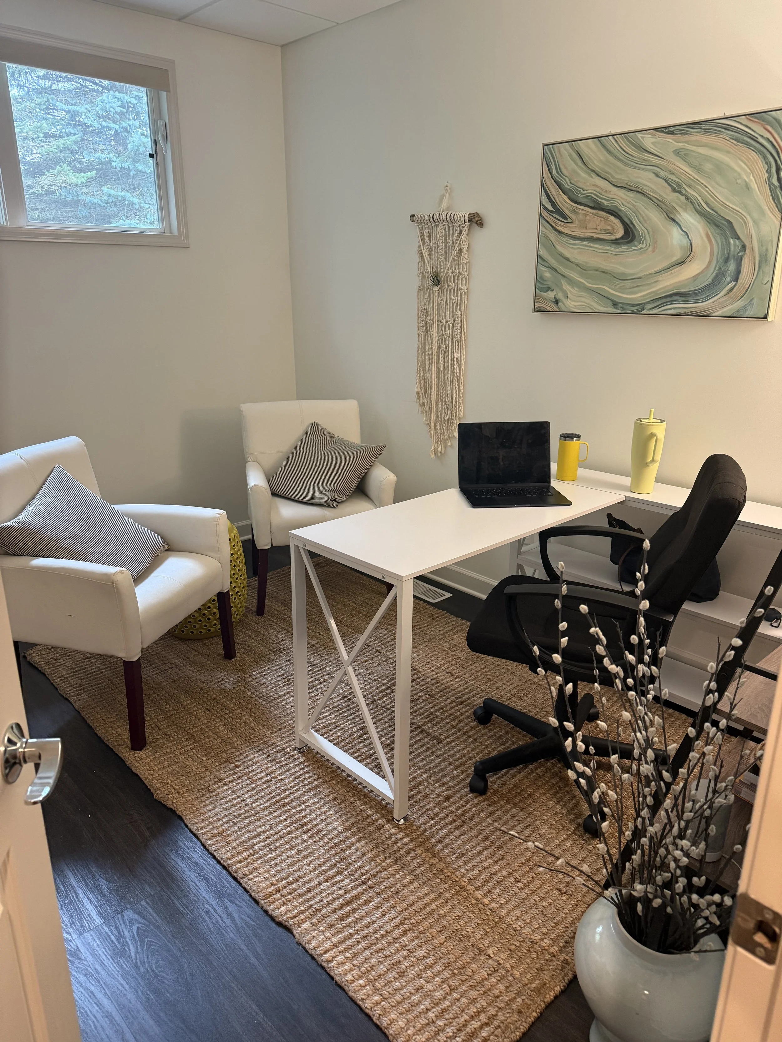 Small home office with white desk, black chair, two white armchairs with gray pillows, a window, a macrame wall hanging, a large abstract art piece, yellow thermos and mug, a laptop, and a petal arrangement in a white vase on the floor.