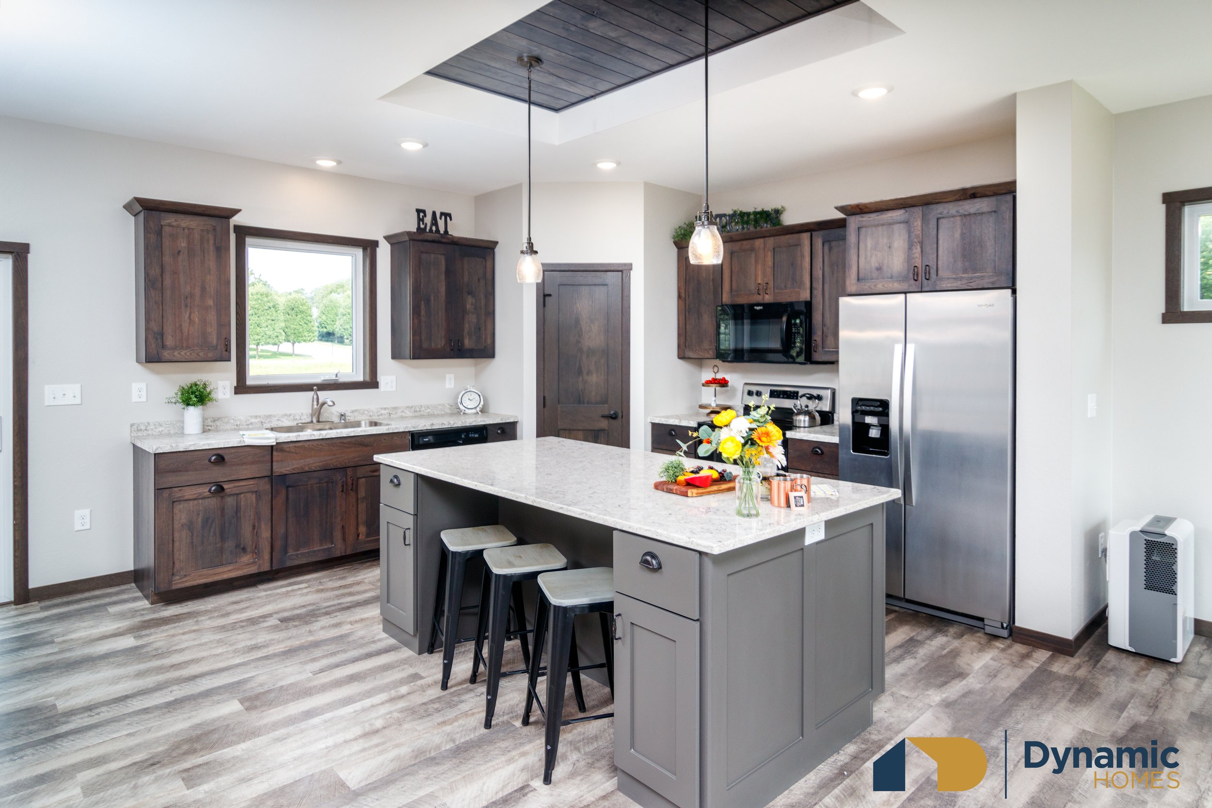 Modern kitchen inside modular home with white walls, wooden cabinets, and a gray kitchen island with three stools, decorated with a bouquet of flowers and some small kitchen items.