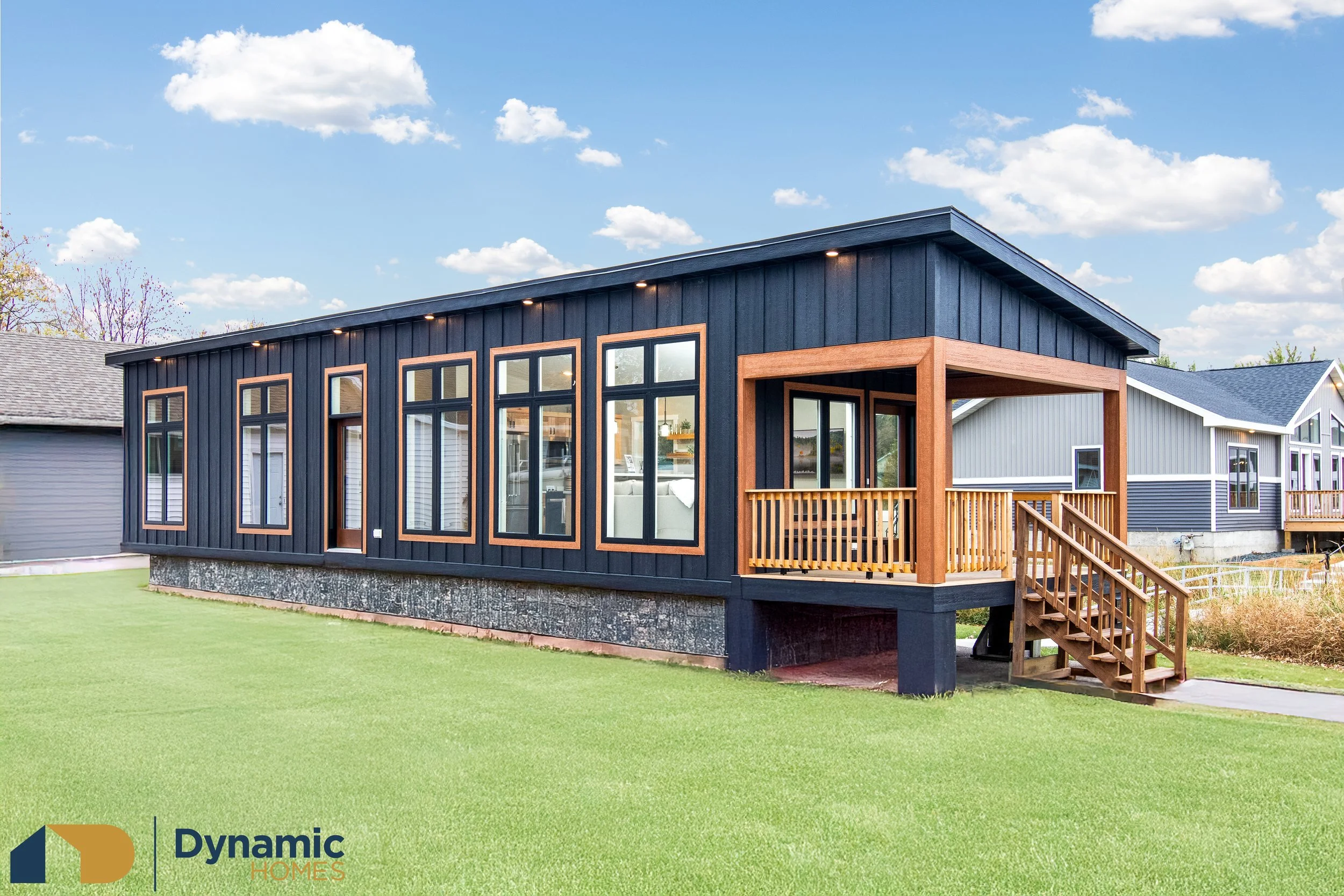 Modular home with dark vertical siding, large windows, a wooden porch, and stairs, set in a green yard under a blue sky with scattered clouds.