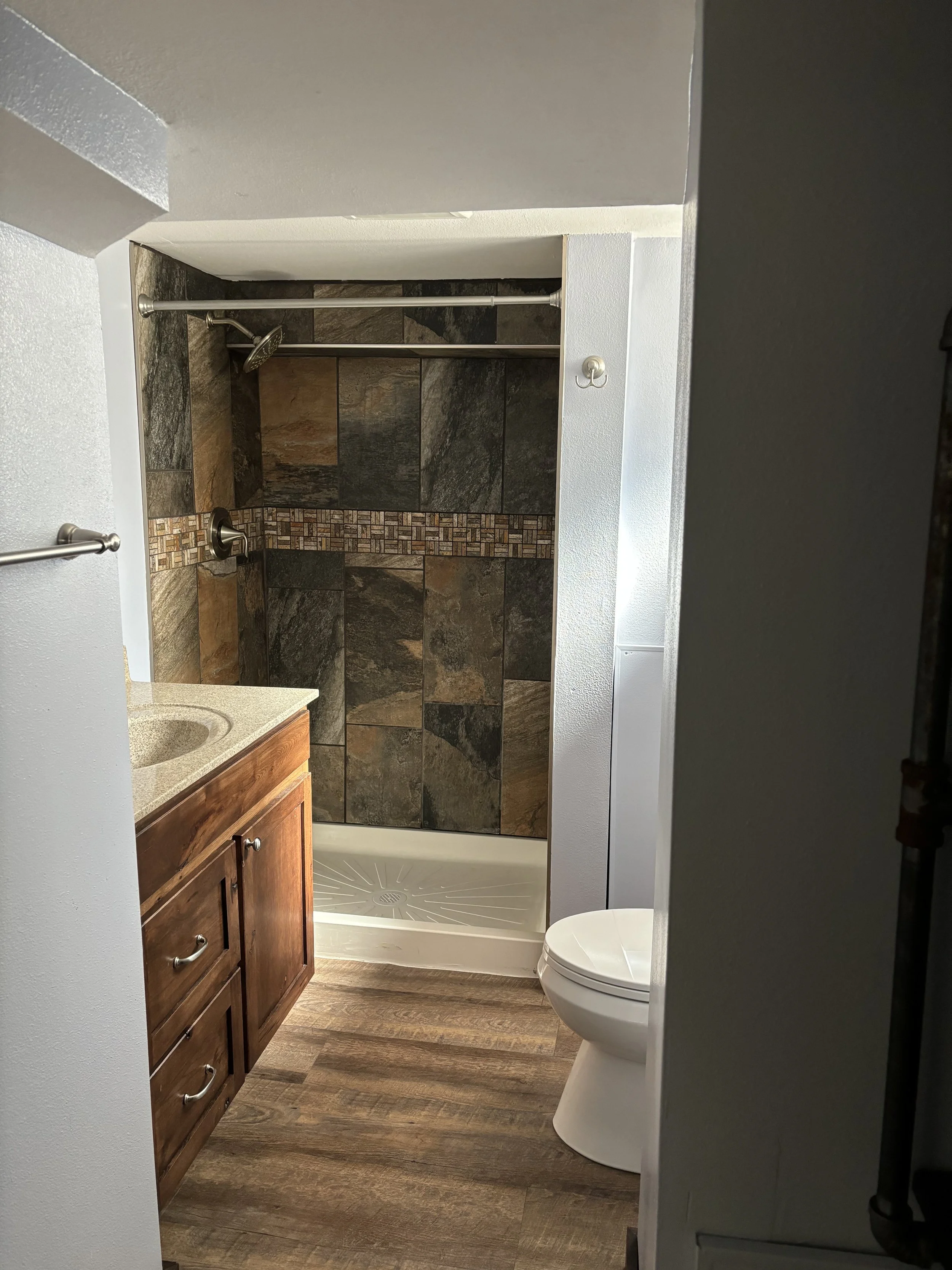 Finished bathroom with a shower, toilet, and wooden vanity with a sink, white walls, and wood-patterned flooring.