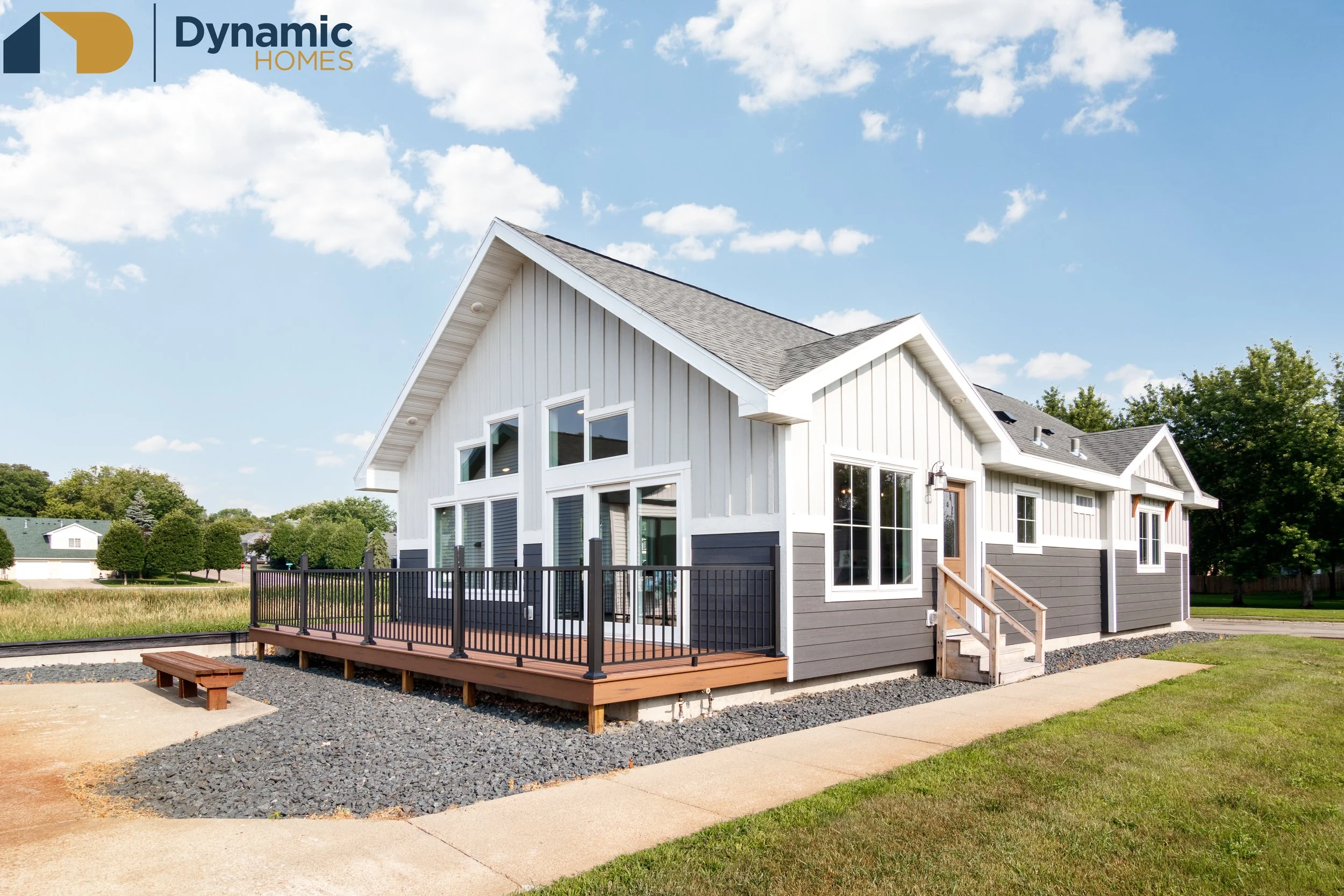 A two-story modular home with white and gray siding, large windows, and a sloped roof, surrounded by a lawn and trees, under a partly cloudy sky.