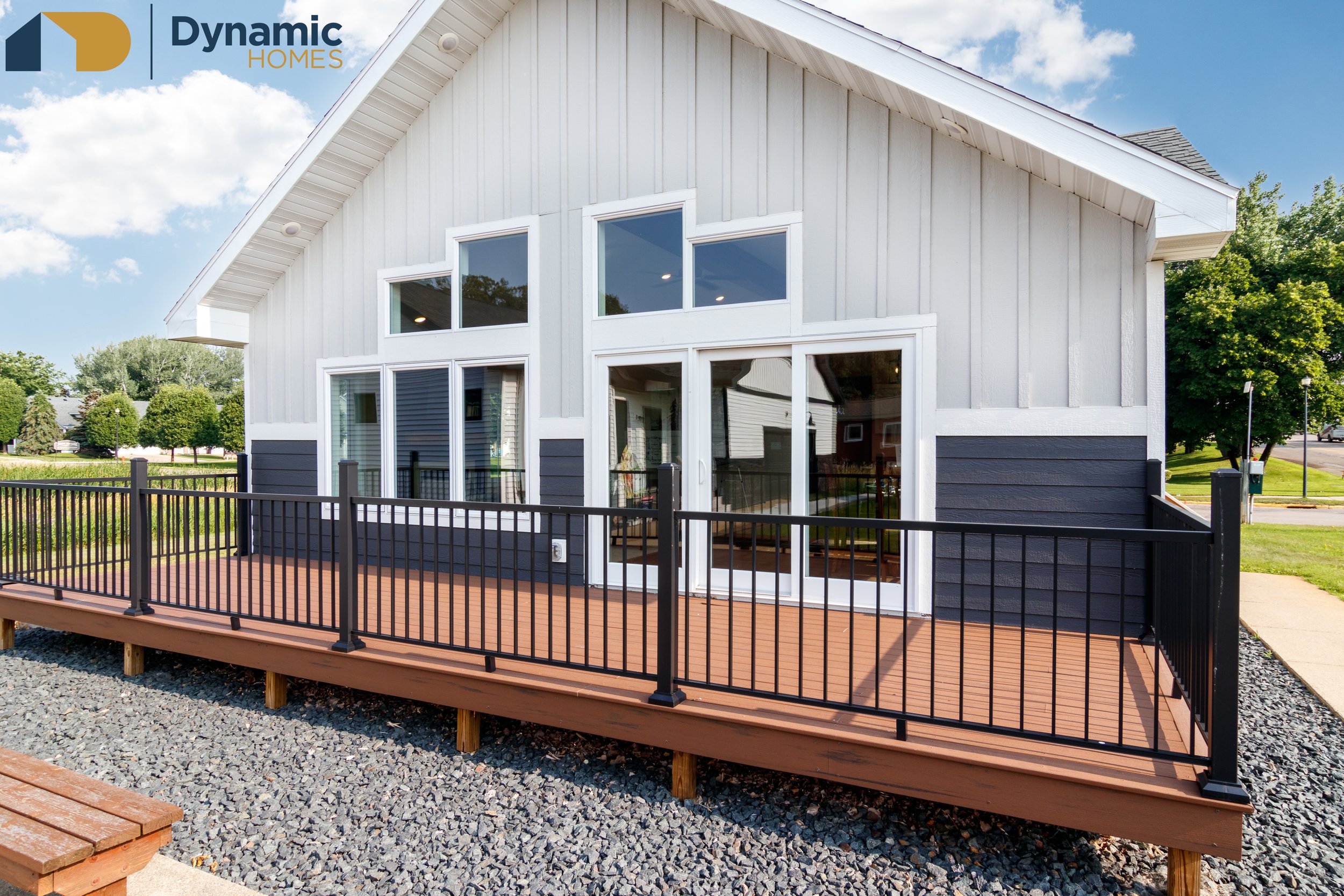 A two-story modular home with a white exterior and a gabled roof, featuring large windows and a sliding glass door that opens to a wooden deck with black metal railing. The house is surrounded by green trees and a well-maintained lawn.