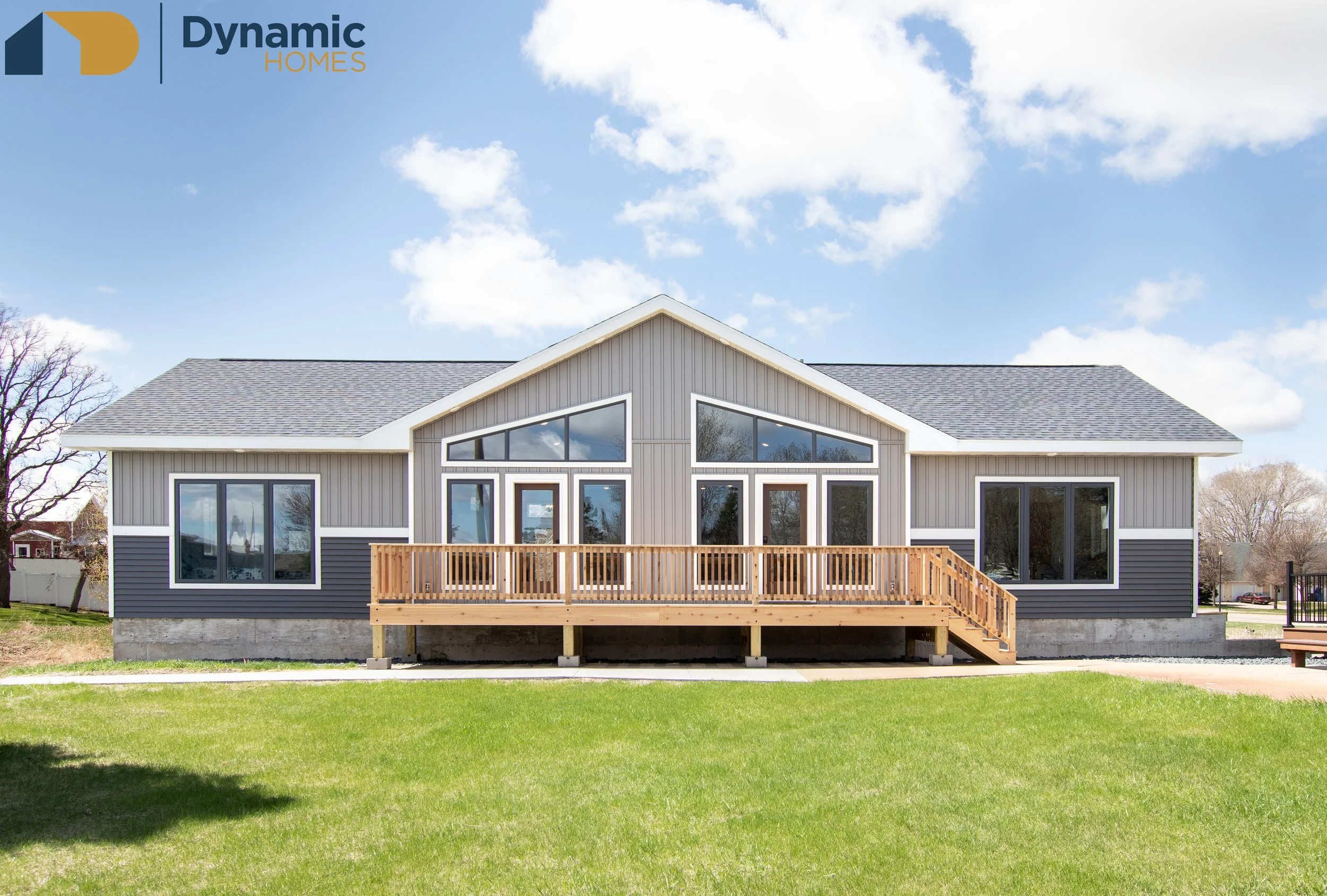 Front view of a gray modular home with large windows, a wooden deck, and a sloped roof, set against a blue sky with scattered clouds and surrounded by green grass and trees.