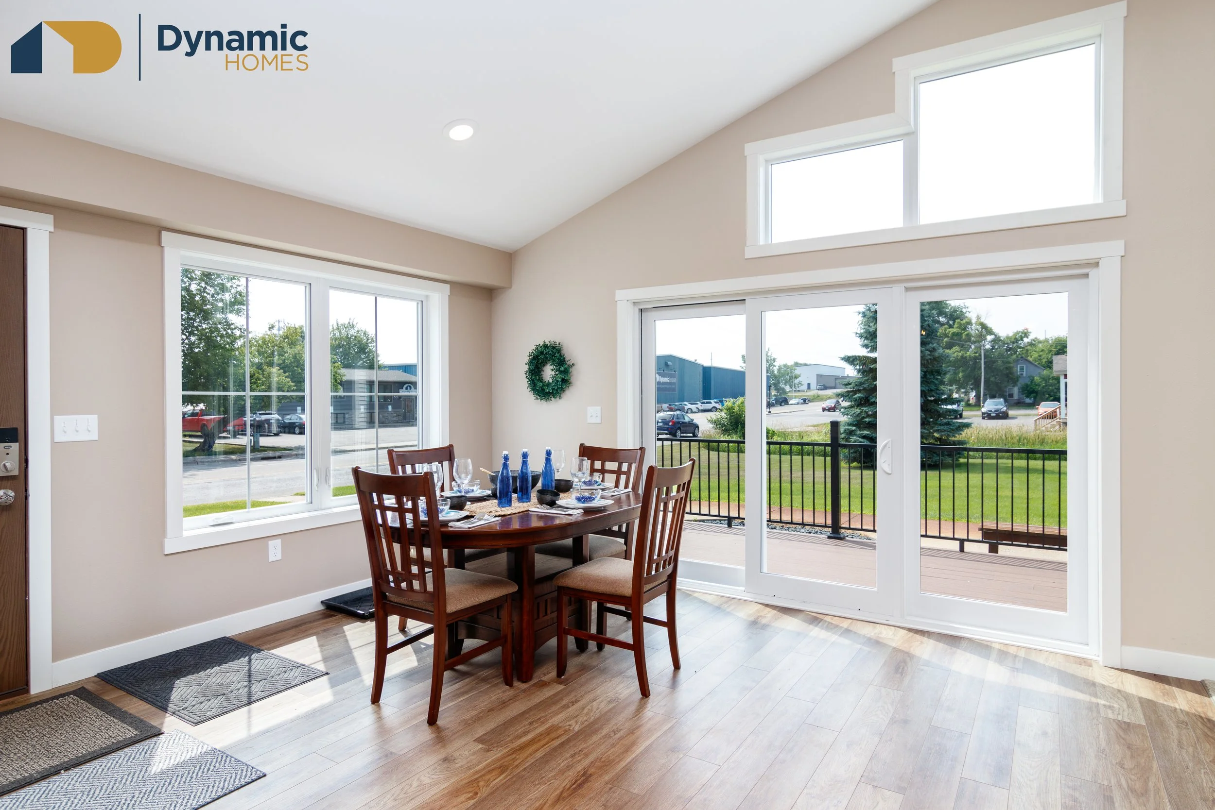 Inside of a modular home with large windows and sliding glass door to a deck, dining table set for a meal, with a green wreath on the wall outside.