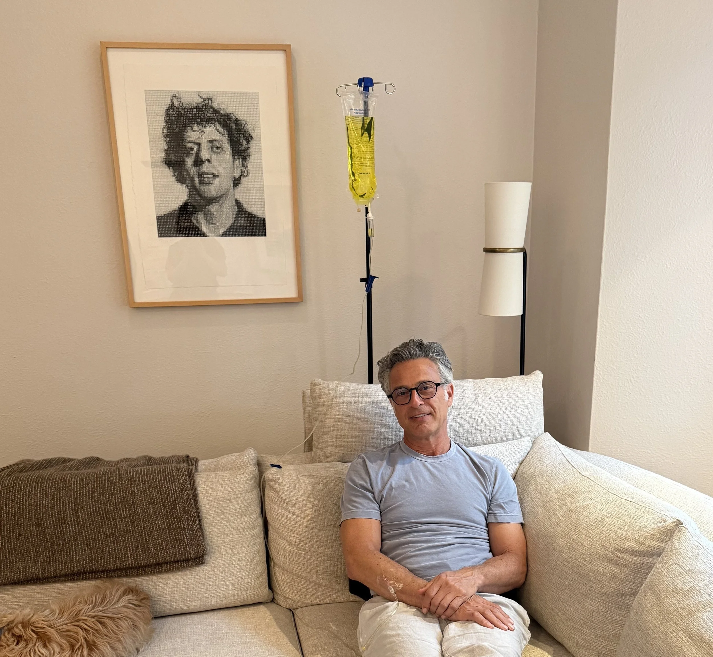 A middle-aged man with glasses and gray hair sitting on a beige sofa in a hospital room, connected to an IV drip, with a framed pixelated portrait of a woman hanging on the wall behind him.