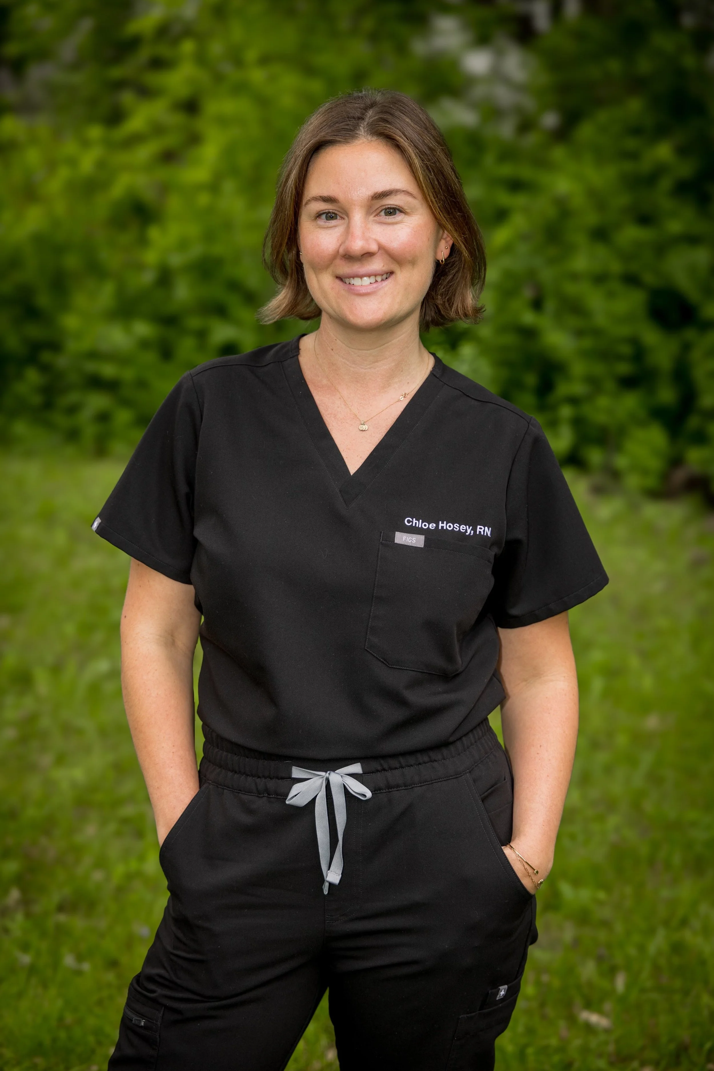 A woman wearing black medical scrubs with the name tag Chloe Hosey, RN, standing outdoors in front of green trees, smiling at the camera.
