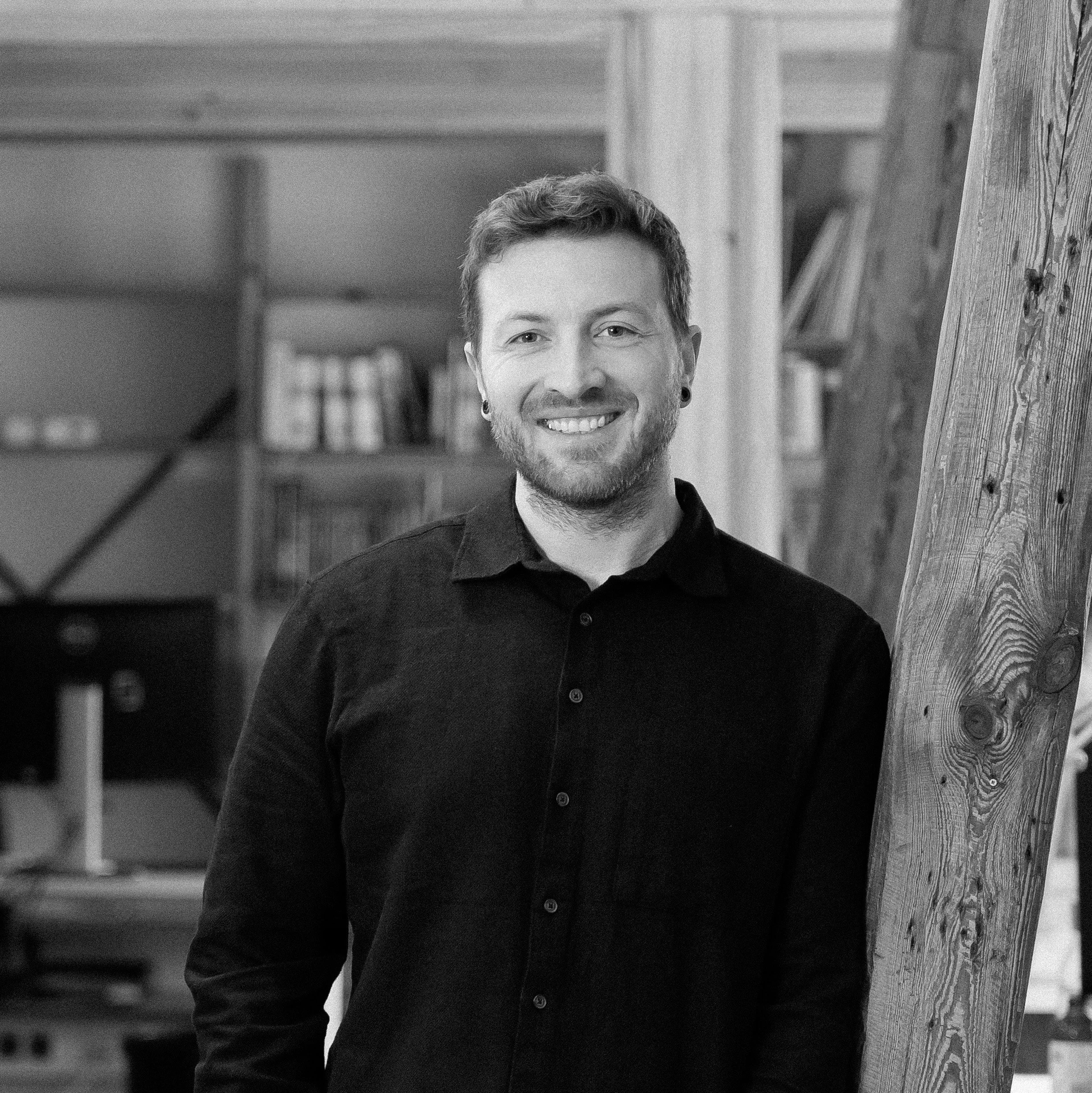 Junge Mann in schwarzem Hemd steht in einem Büro mit Holzwänden und -stützen, lächelnd, mit Händen in den Taschen.