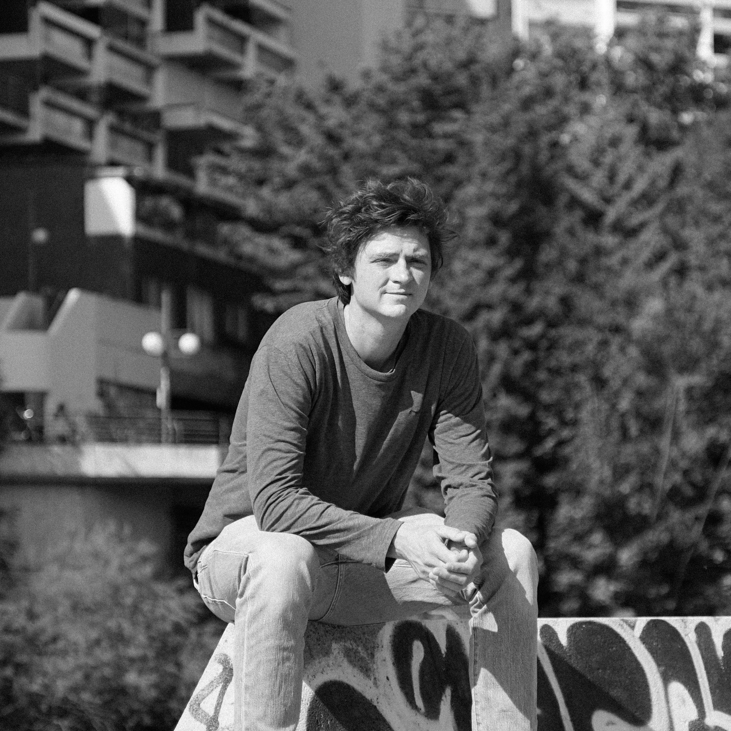 Junge sitzt auf einer grauen, mit Graffiti versehenen Bauruine im Freien, im Hintergrund sind grüne Bäume und moderne Hochhäuser.