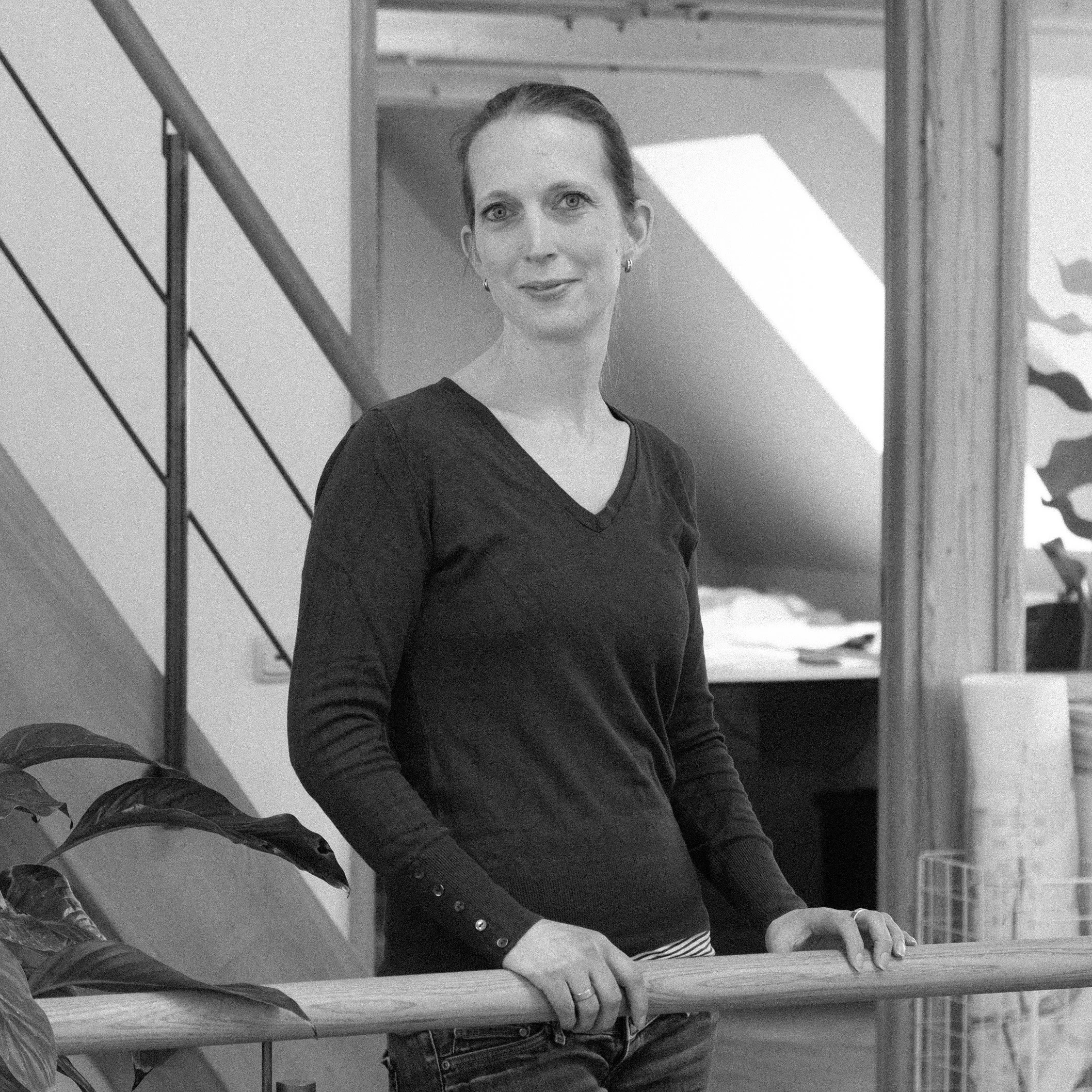 Eine junge Frau mit grauen Hosen, schwarzen Pullover und Brille steht in einer Türöffnung im Eingangsbereich eines Hauses, mit einem Garten im Hintergrund.