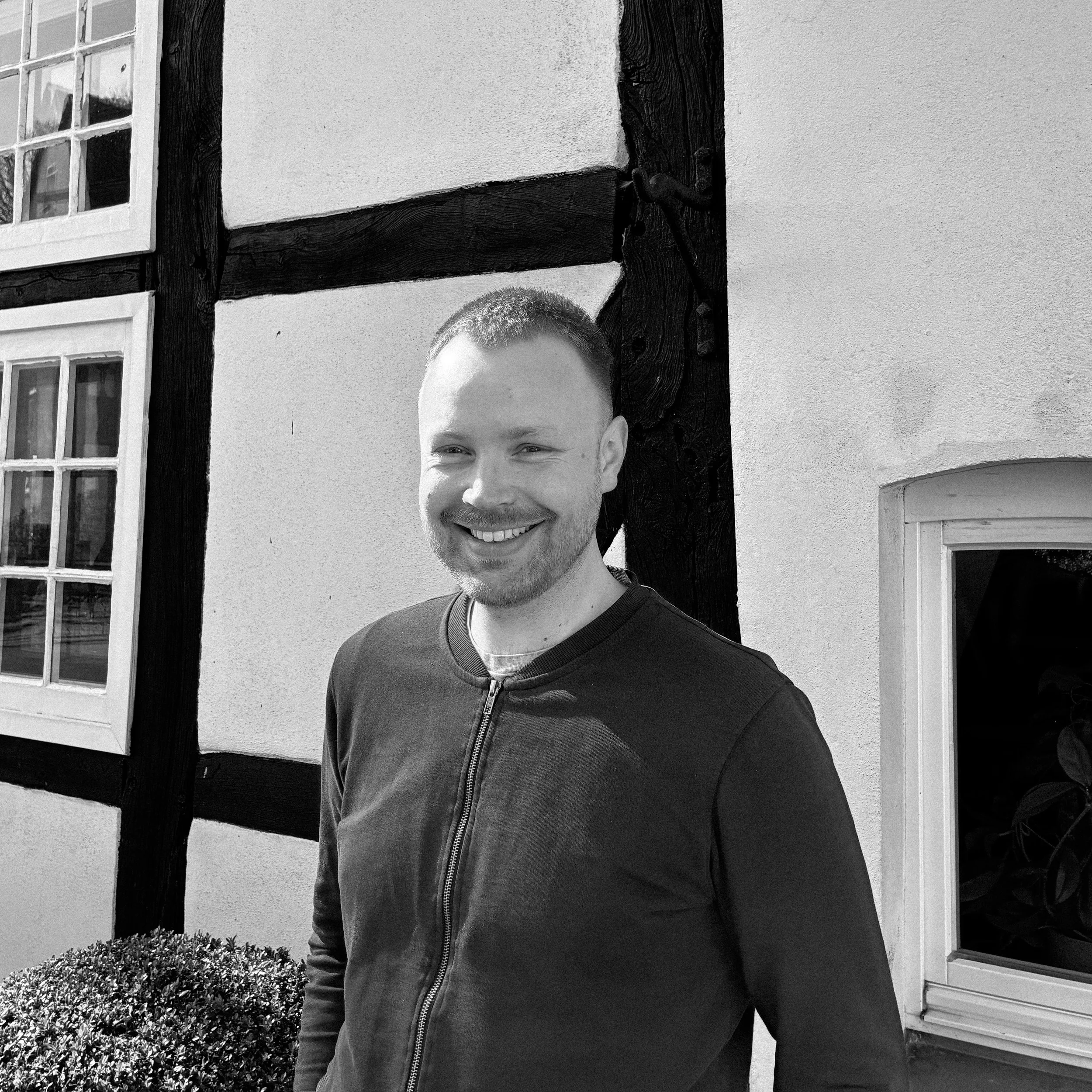 Junger Mann mit Brille, blondes Haar, im weißen T-Shirt mit Schriftzug, lächelnd, im Freien vor grüner Natur.