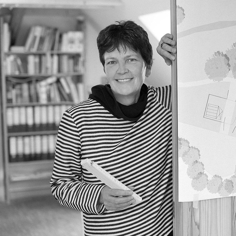 Frau mit kurzen braunen Haaren, schwarz-weiß gestreiftem Pullover, hält ein Notizbuch, steht vor einem Wand mit Landkarte, im Hintergrund Bücherregale.