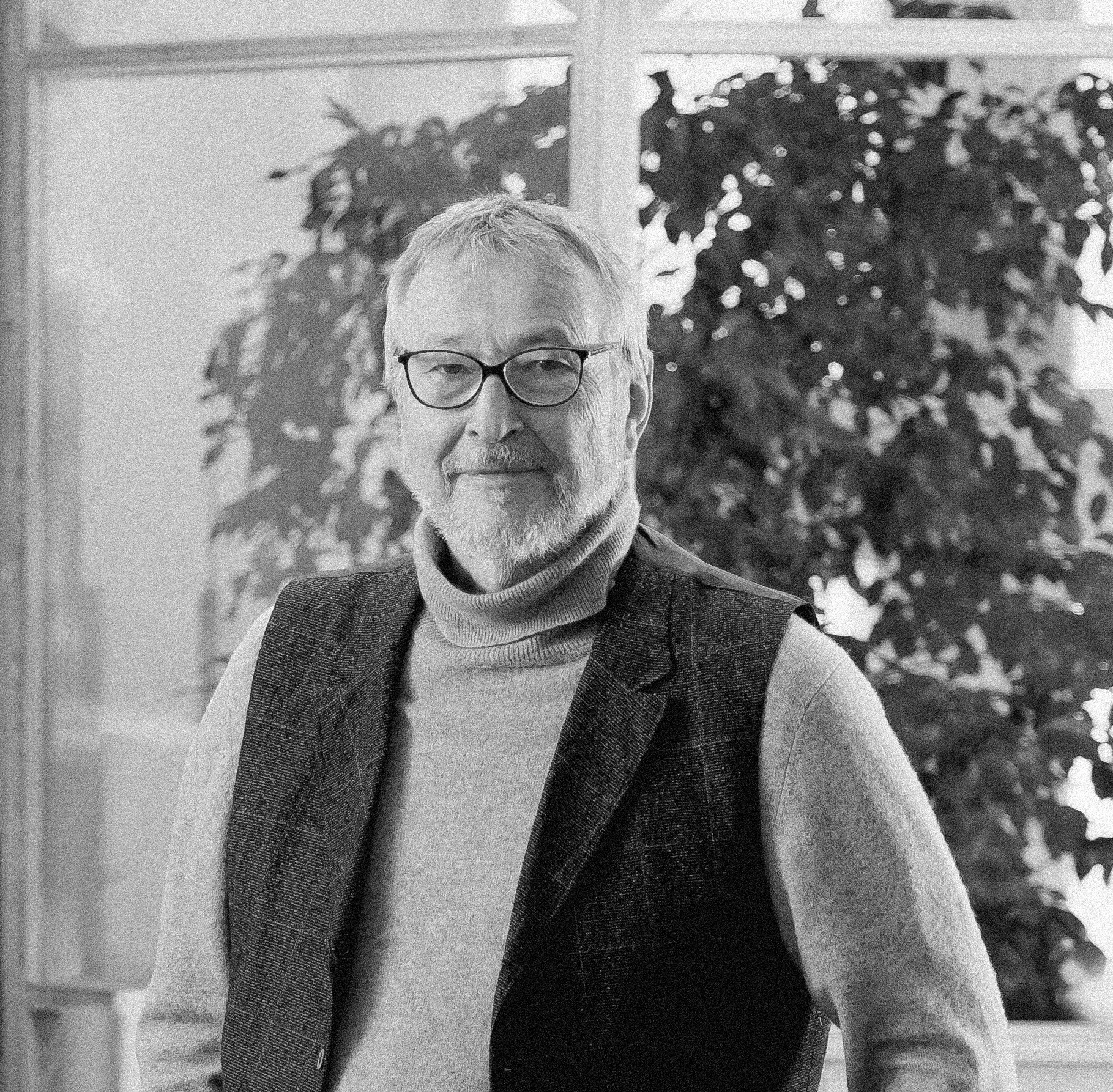 Ein älterer Mann mit Brille, grauem Bart, grauen Haaren, trägt ein graues Turtleneck und eine dunkle Weste, steht in einem modernen Büro und lächelt.