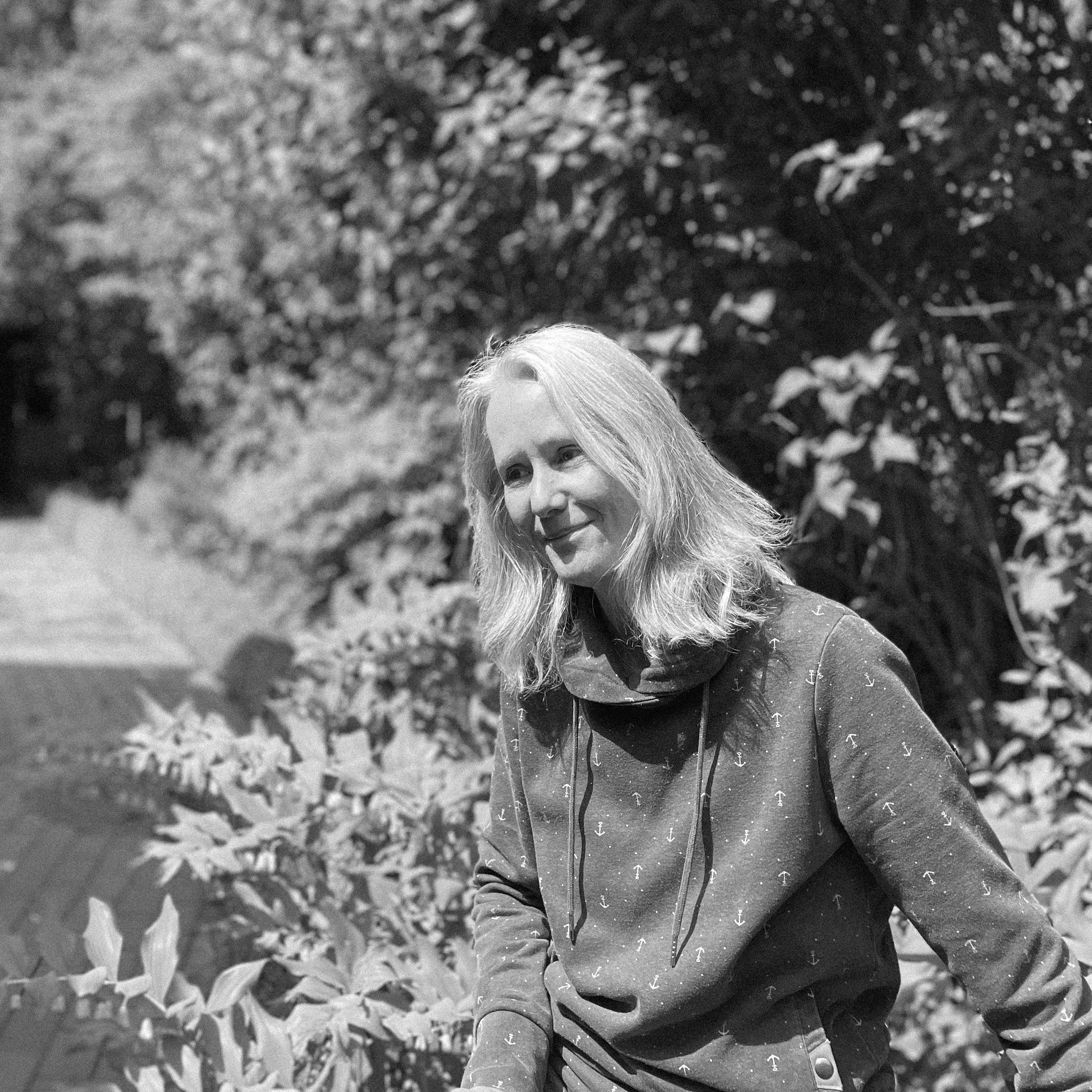 Junge Frau mit blonder, schulterlanger Haar trägt grauen Hoodie mit Anker-Design, steht im Grünen, lächelt, im Hintergrund ein Garten mit Bäumen und einer Gartenbank.