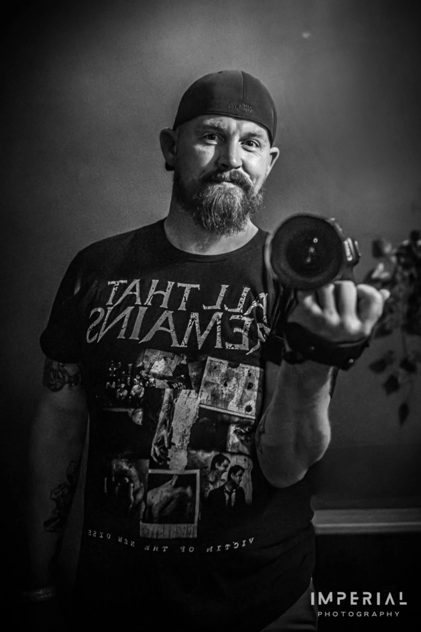 A man with a beard and mustache taking a black and white selfie in a mirror, wearing a backwards cap and a graphic T-shirt. He is holding a camera with a strap, and the background appears to be a room with plain walls and a plant.