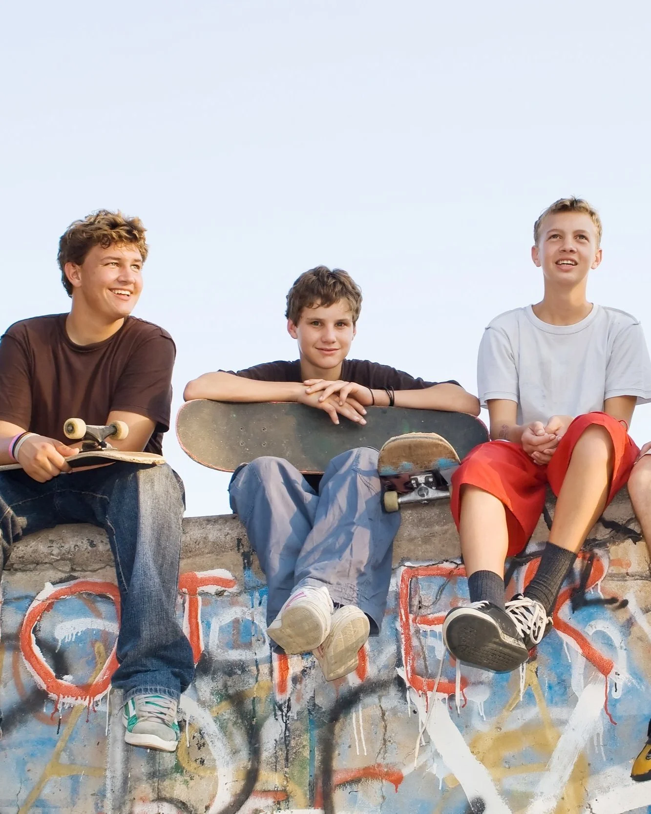 Three teenage boys sitting on a graffiti-covered wall at a skate park, with one holding a skateboard and another holding a longboard, all smiling and wearing casual clothes.