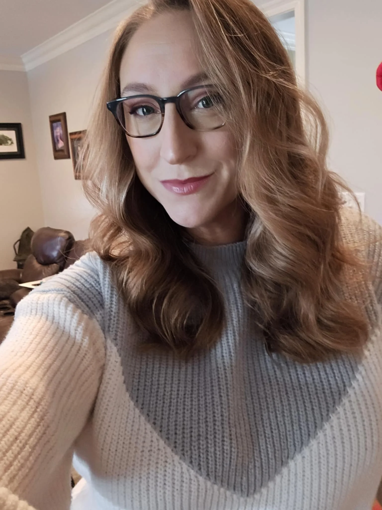A woman with wavy, reddish-brown hair and glasses, smiling in a beige sweater, takes a selfie in a living room.