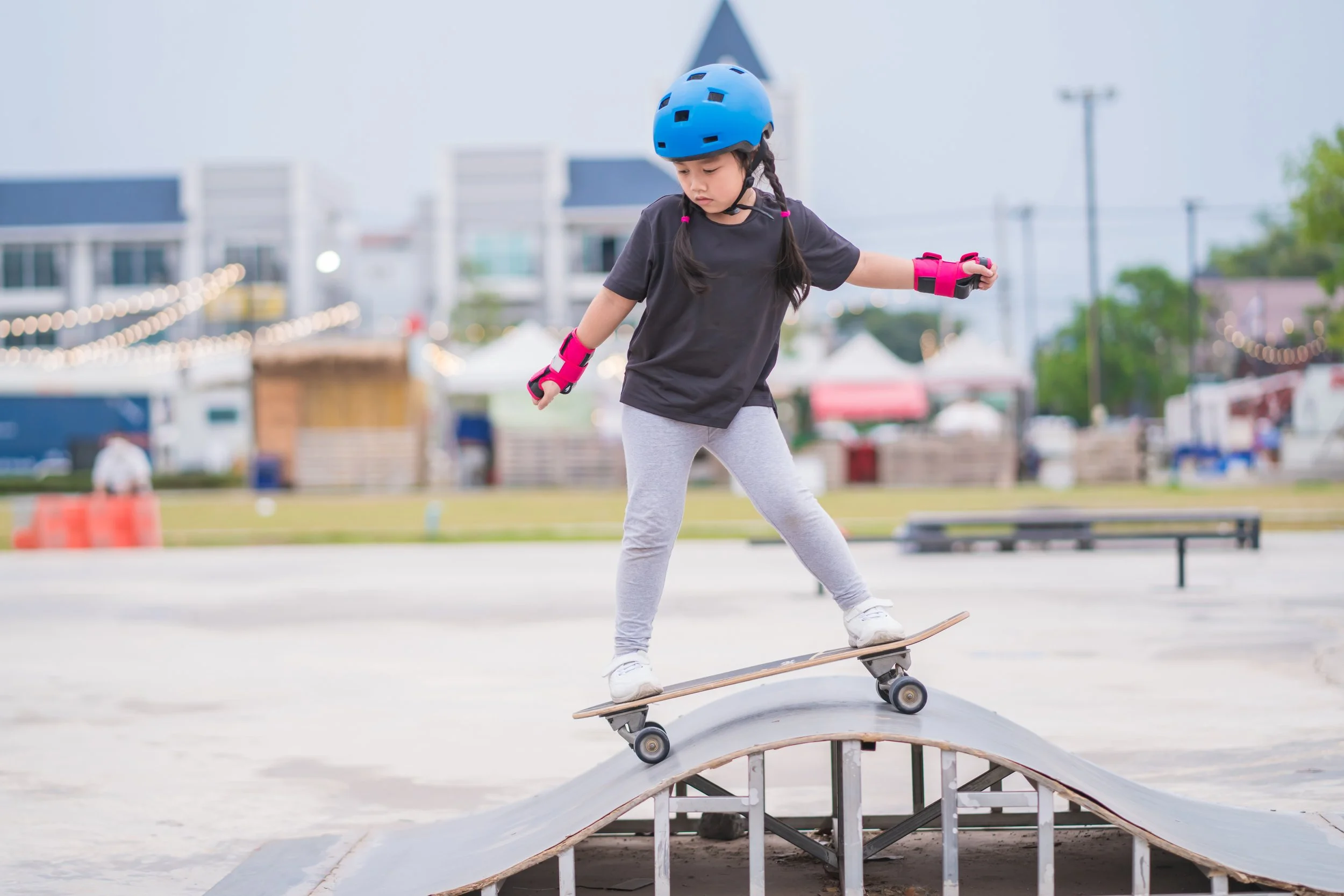 A young girl wearing a blue helmet and pink protective gear skateboarding on a small ramp at an outdoor skate park.