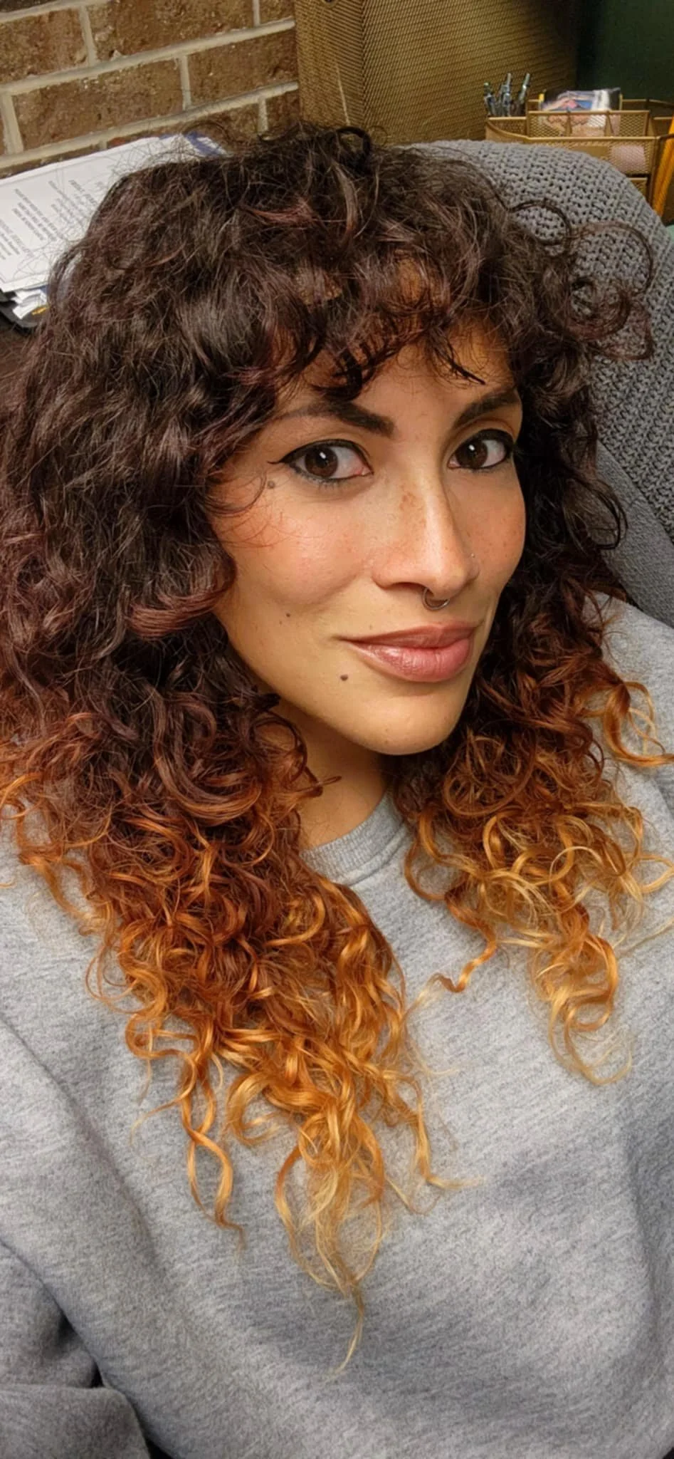 A woman with curly, two-toned hair, makeup, and a septum piercing, sitting on a gray textured chair in a room with brick walls and office supplies in the background.
