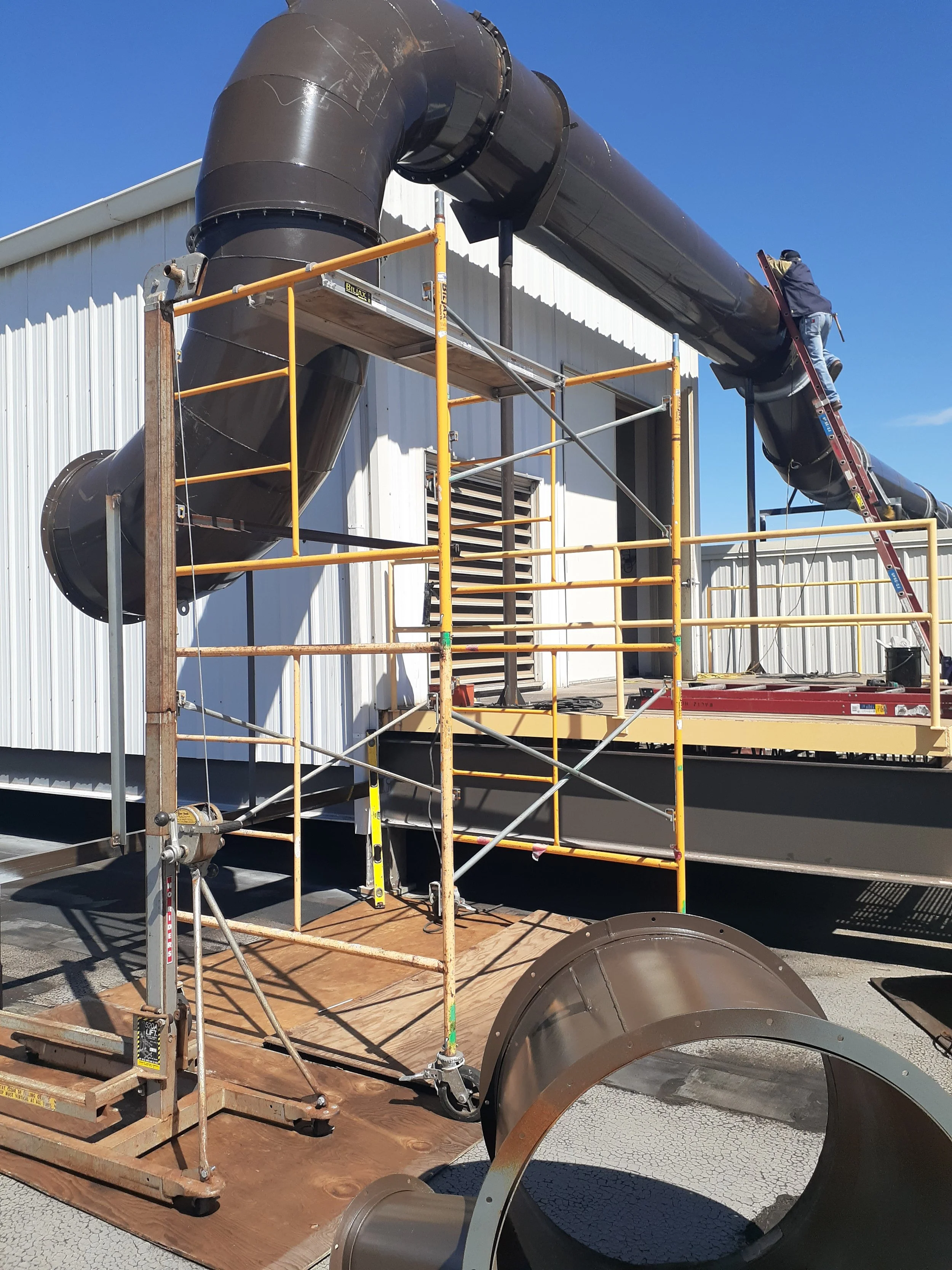 A worker repairs or installs large black industrial pipes on the side of a white metal building, standing on a yellow scaffolding and a red ladder, under a clear blue sky.
