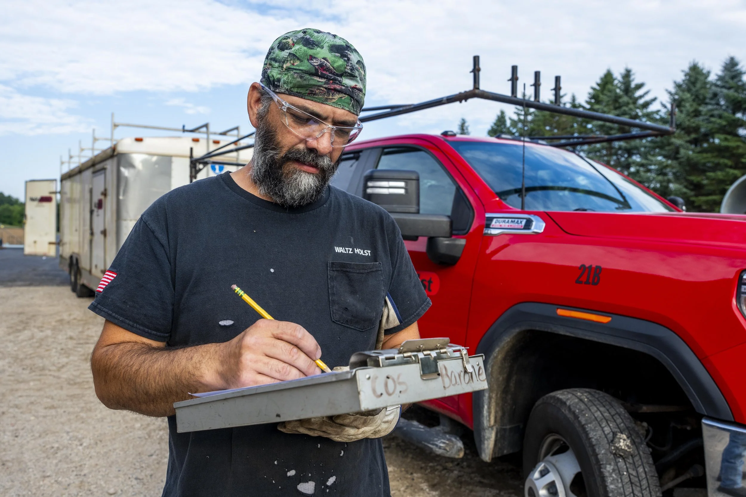 A man with a beard, glasses, and a camouflage cap writes on a clipboard, standing near a red truck with a ladder on top and a trailer in the background.