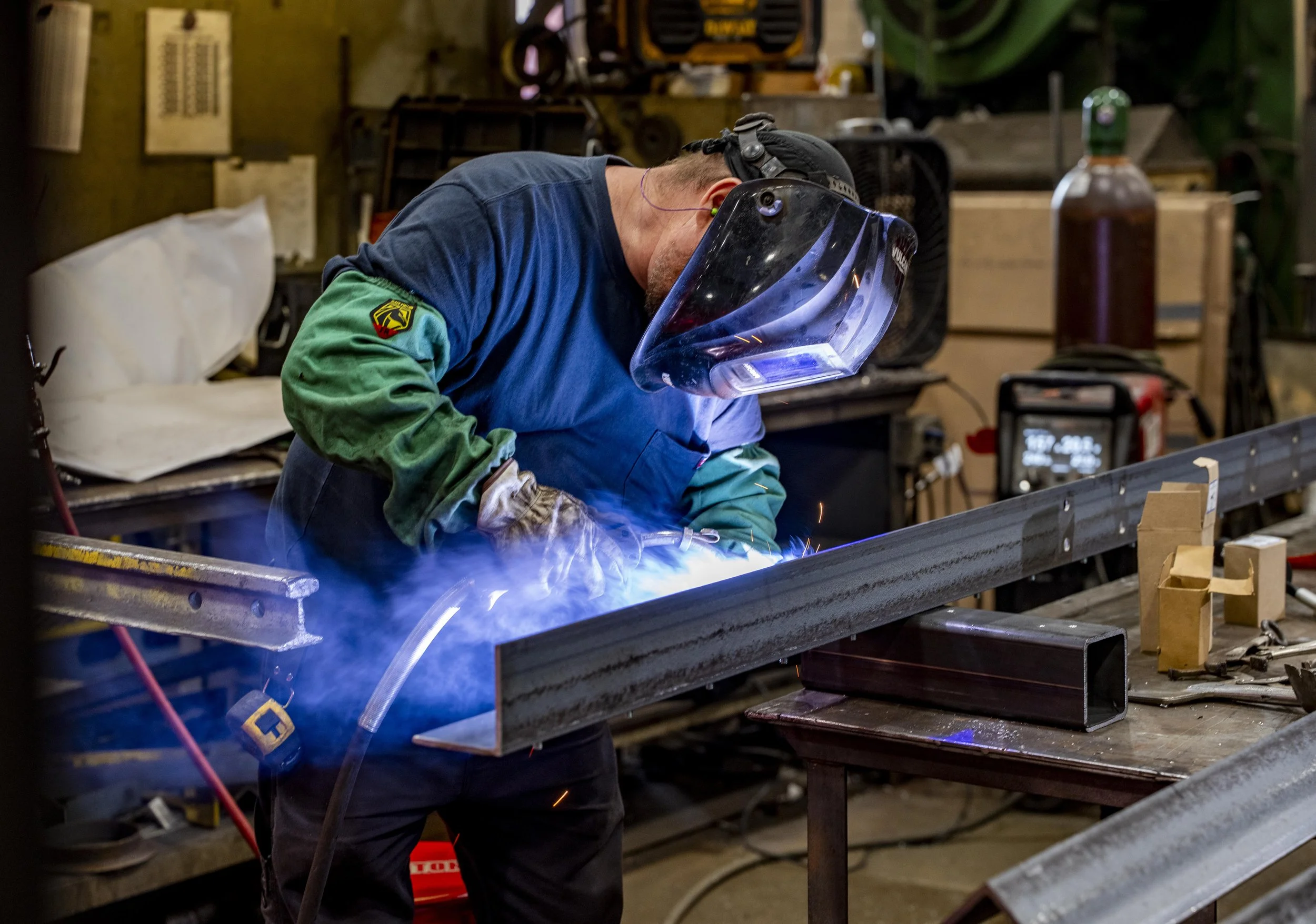 A welder wearing protective gear, including a helmet and gloves, is welding a square metal tube in a workshop with various tools and equipment.