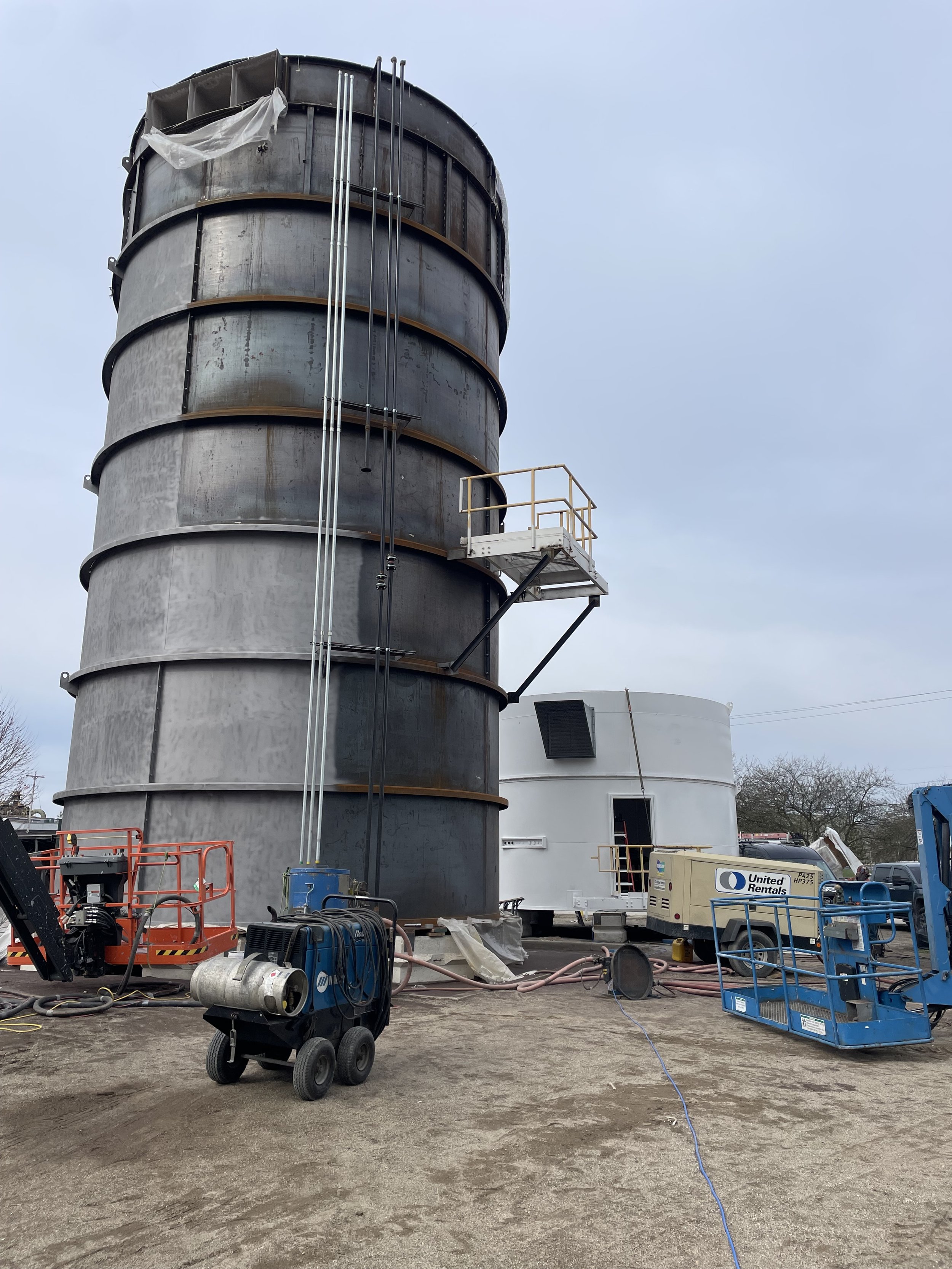 Construction site with a large, partially assembled dust collector system, a white mobile building, and construction equipment, including a blue air compressor and a blue lift platform.