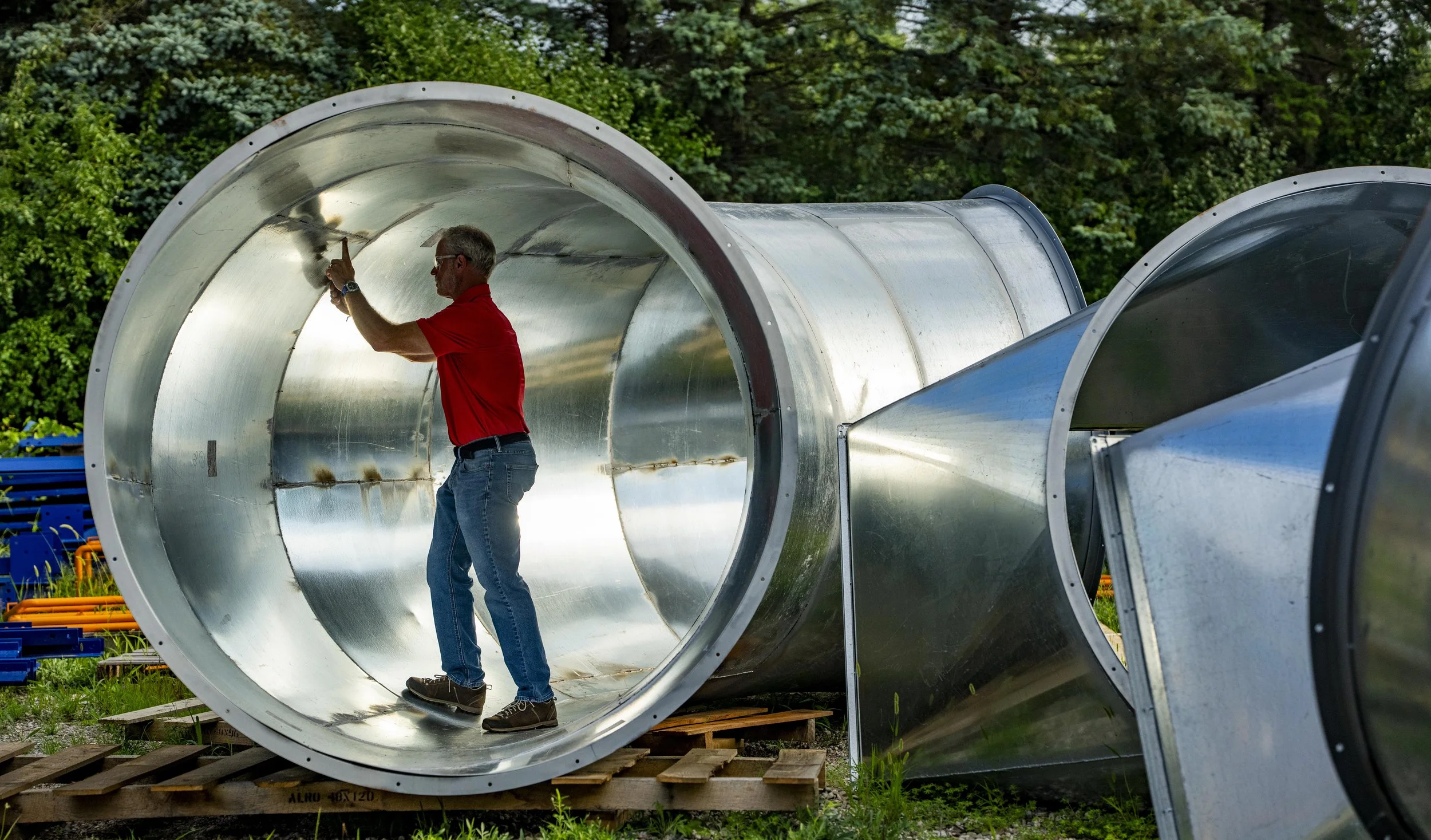 A man wearing glasses, a red shirt, and jeans is inspecting and cleaning the inside of a large metallic ventilation duct outdoors. Several similar ducts are lying on the ground nearby, with trees in the background.
