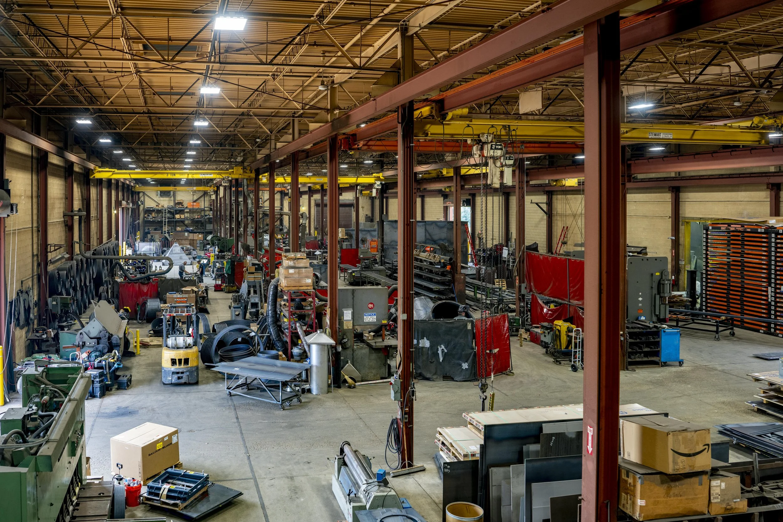 Interior view of a large industrial workshop with high ceiling, metal beams, and bright overhead lighting, filled with various machinery, tools, and equipment.